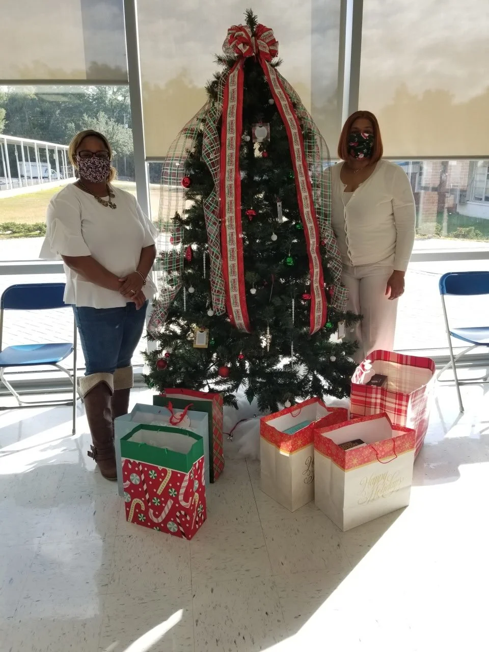 Two women standing on either side of a decorated Christmas tree with gift bags at its base, inside a room with large windows and blinds.