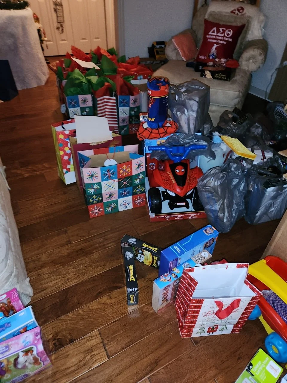 Living room with Christmas gifts, including gift bags, wrapped presents, a Spiderman ride-on toy, and Christmas-themed items on a wooden floor.
