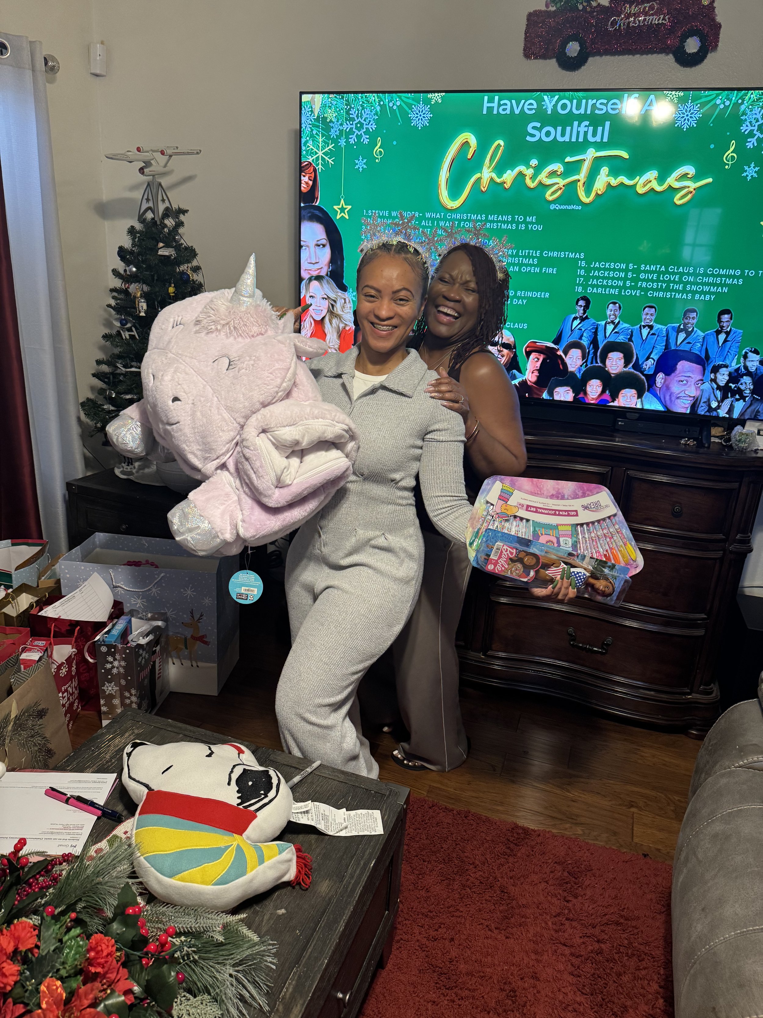 Two women celebrating Christmas in a living room with a TV in the background. One woman is holding a large plush unicorn toy, and the other woman is wearing a silver tiara. The room has Christmas decorations, including a small Christmas tree with ornaments, wrapped presents, and a table with holiday-themed items. The TV screen displays a festive Christmas program.