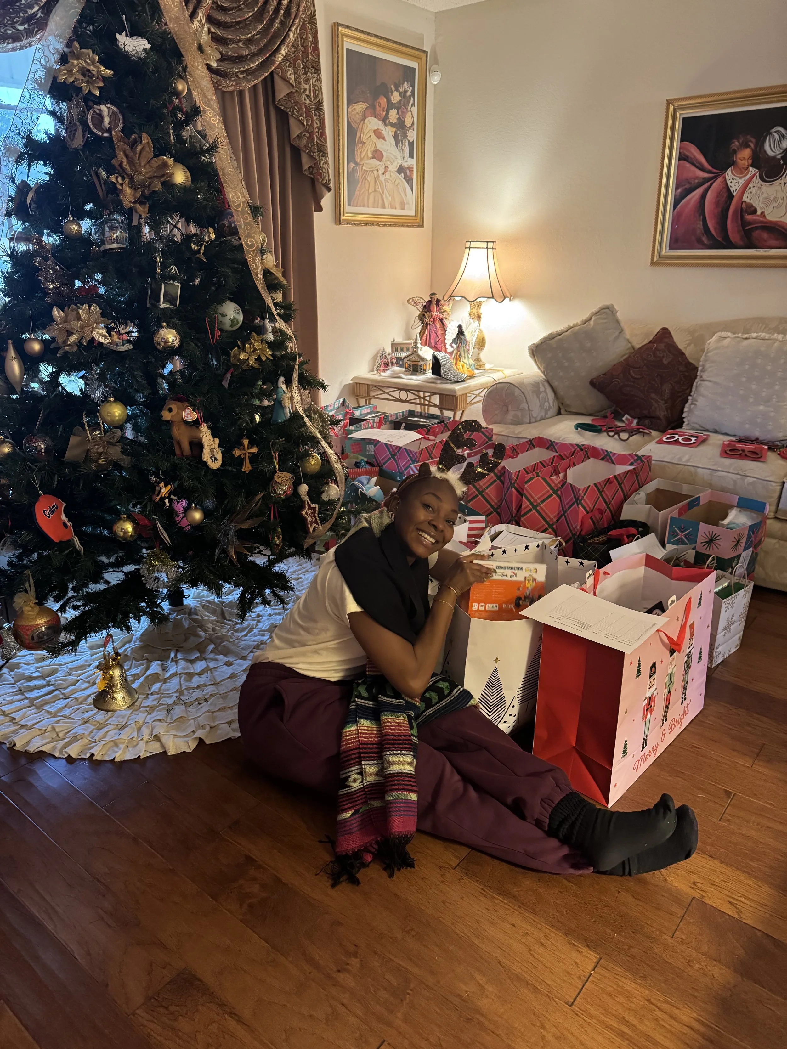 A woman smiling, sitting on the floor in front of a decorated Christmas tree and surrounded by gift bags. She is wearing reindeer antlers and a striped scarf.