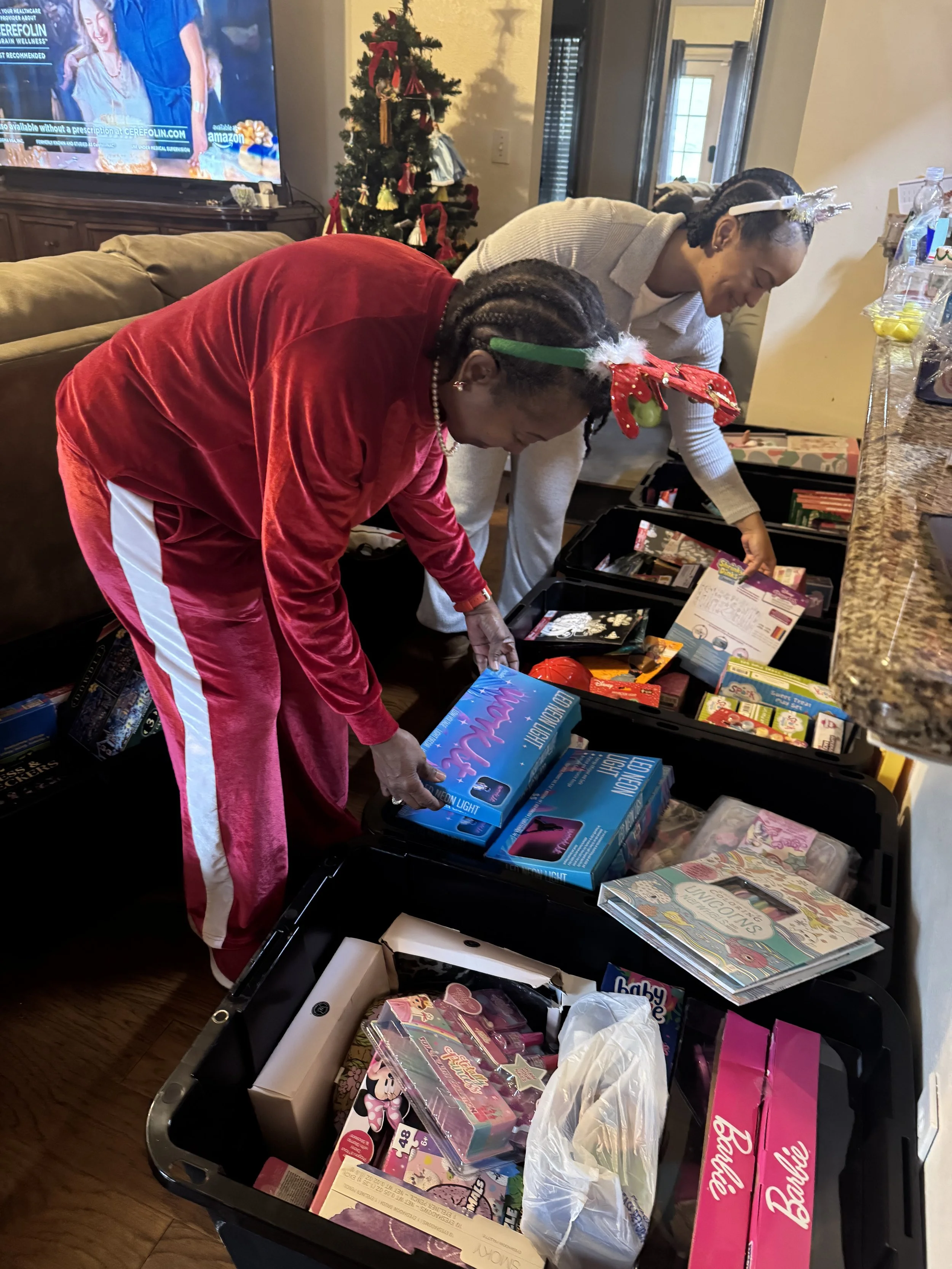 Two women are bent over a laundry basket filled with various toys and gift boxes, in a room decorated for Christmas with a Christmas tree in the background, a television, and a granite countertop with water bottles and treats.