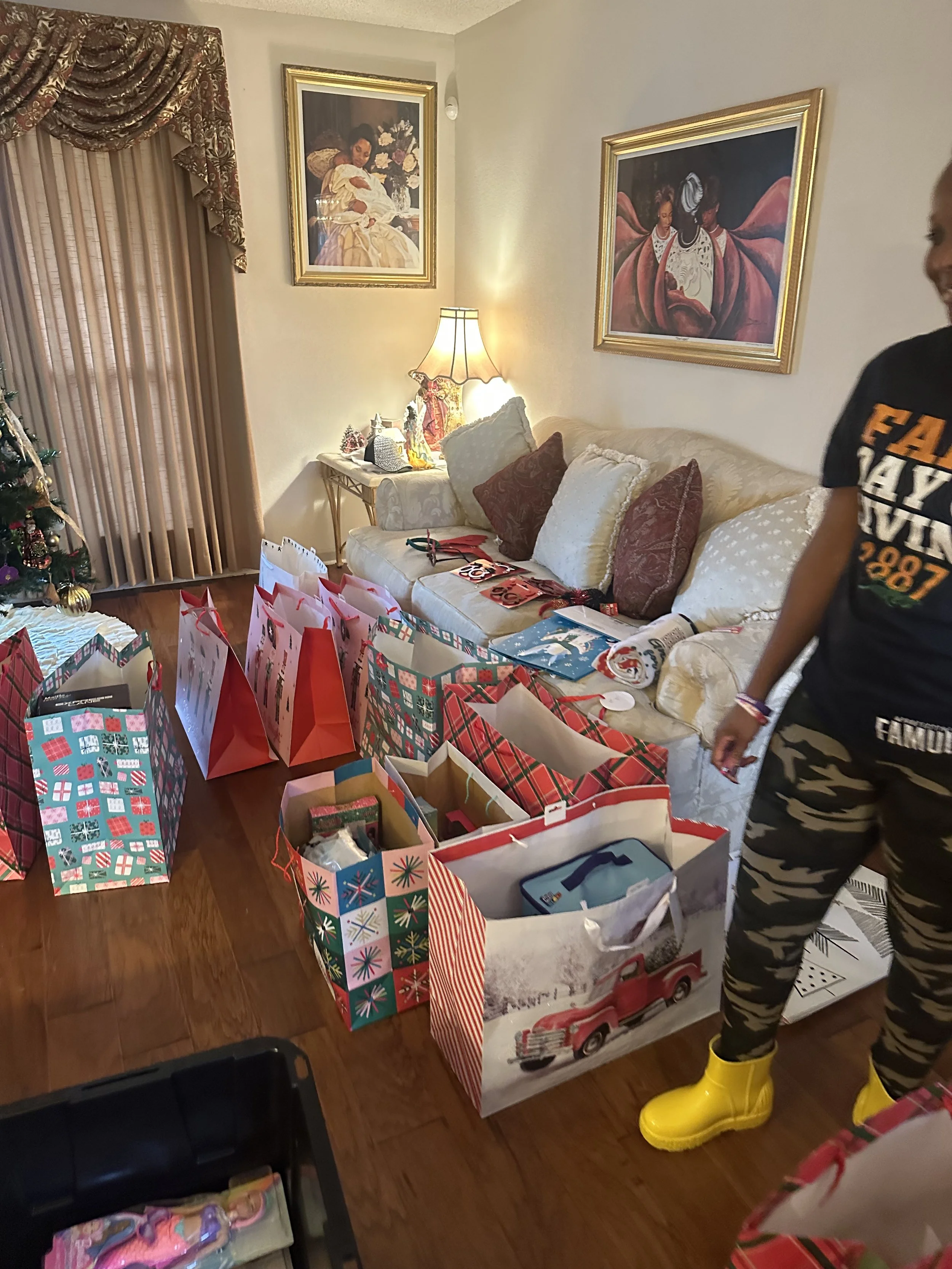 A cozy living room decorated for Christmas with a tree, wrapped gifts on a table, and framed art on the walls. A person wearing yellow rain boots and camouflage pants is standing in front of the gifts.