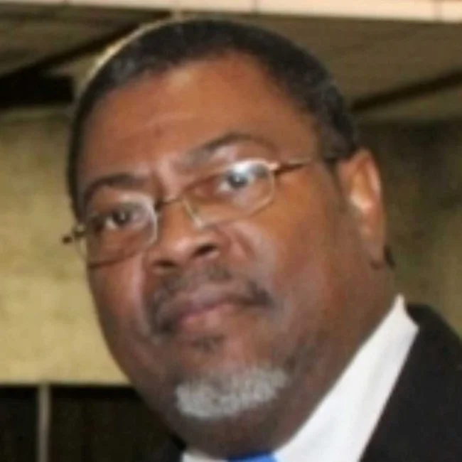 Close-up of a man wearing glasses, with a dark beard and mustache, dressed in a suit with a white shirt and tie.