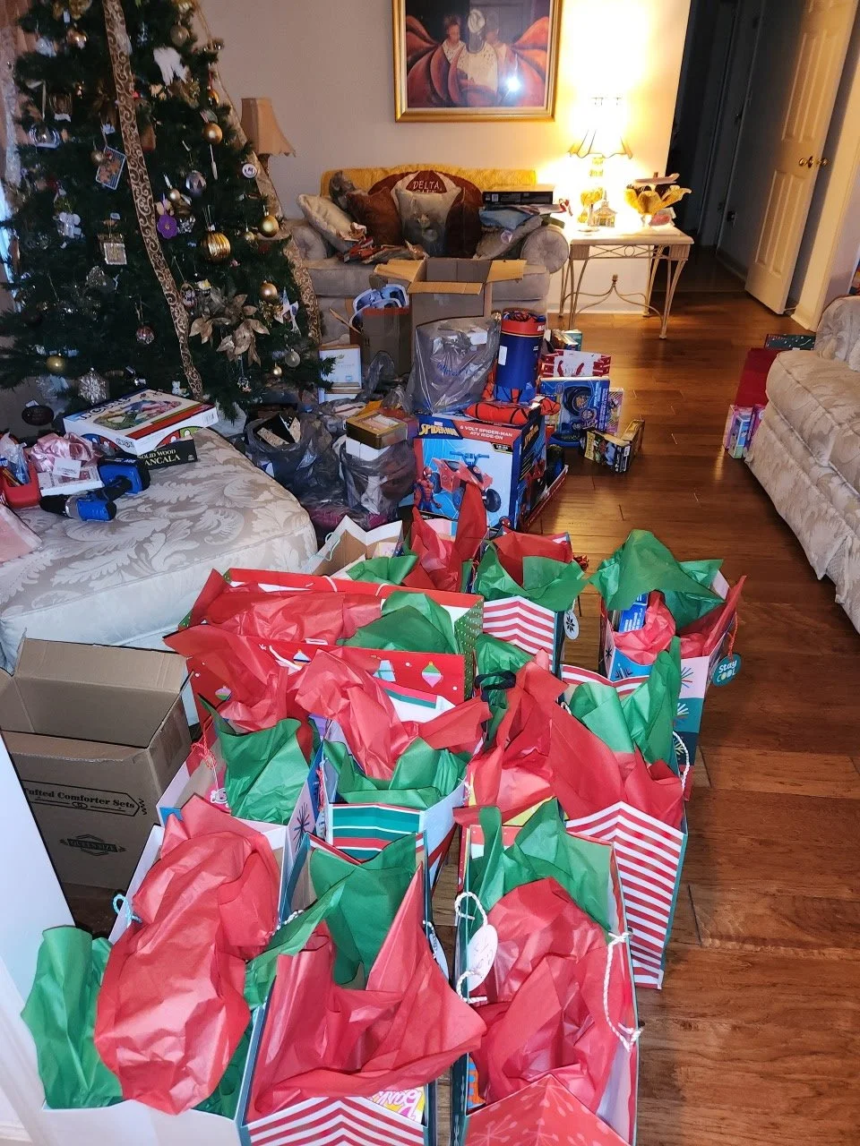 Living room decorated for Christmas with a large decorated Christmas tree, wrapped gifts, toy boxes, and a couch with many pillows.