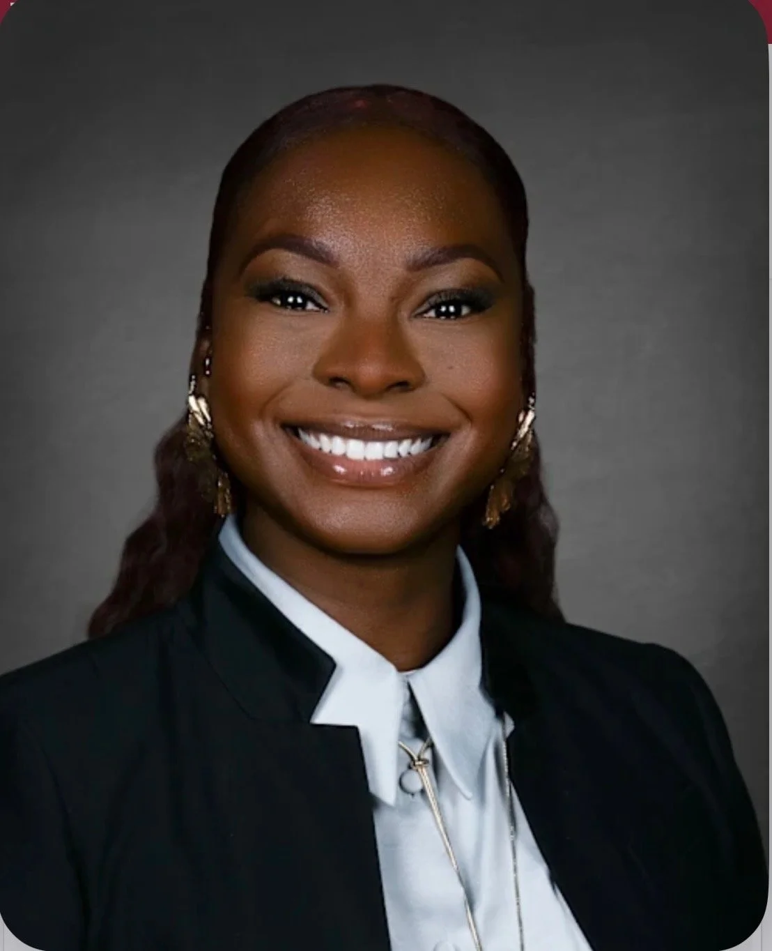 Professional headshot of a smiling African American woman with earrings, wearing a black blazer and white shirt, against a gray background.