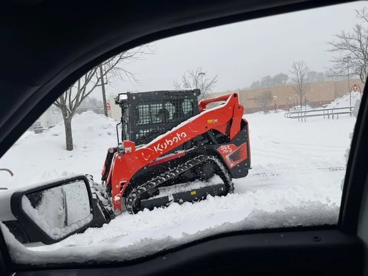 A Kubota snow tractor clearing snow in a parking lot, seen from inside a vehicle during winter.