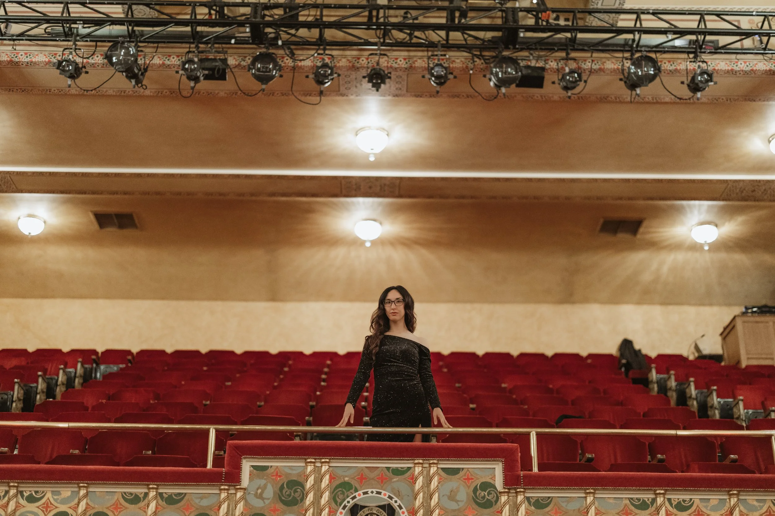 A woman with long hair and glasses standing in the center of an empty theater with red seats and ornate decor. The ceiling has several bright lights and stage equipment hanging from above.