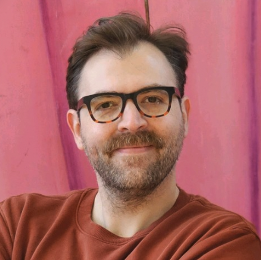 A man with glasses and a beard wearing a brown shirt, smiling against a pink background.