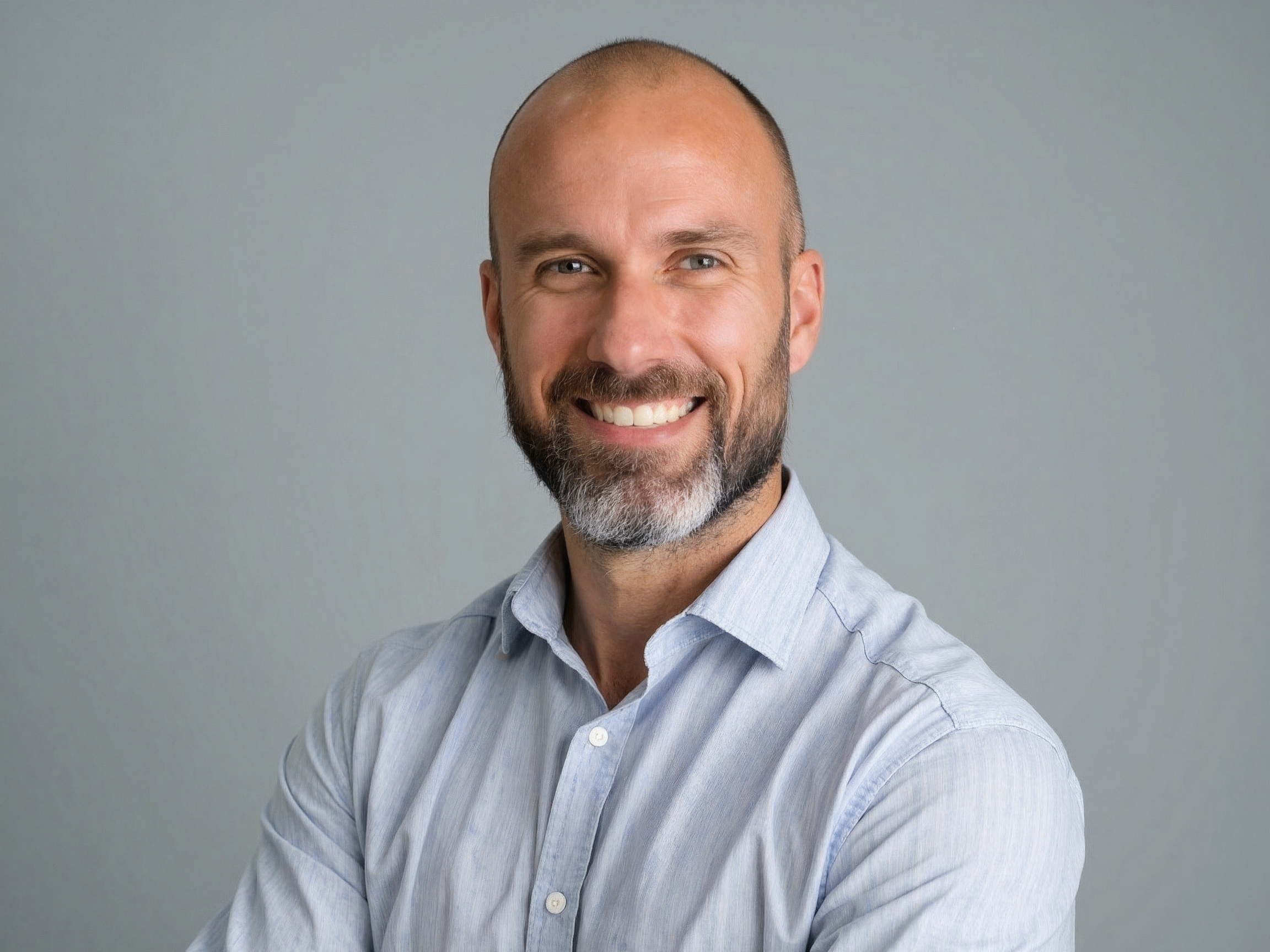 A smiling man with a beard, dressed in a light blue button-up shirt, against a plain gray background.