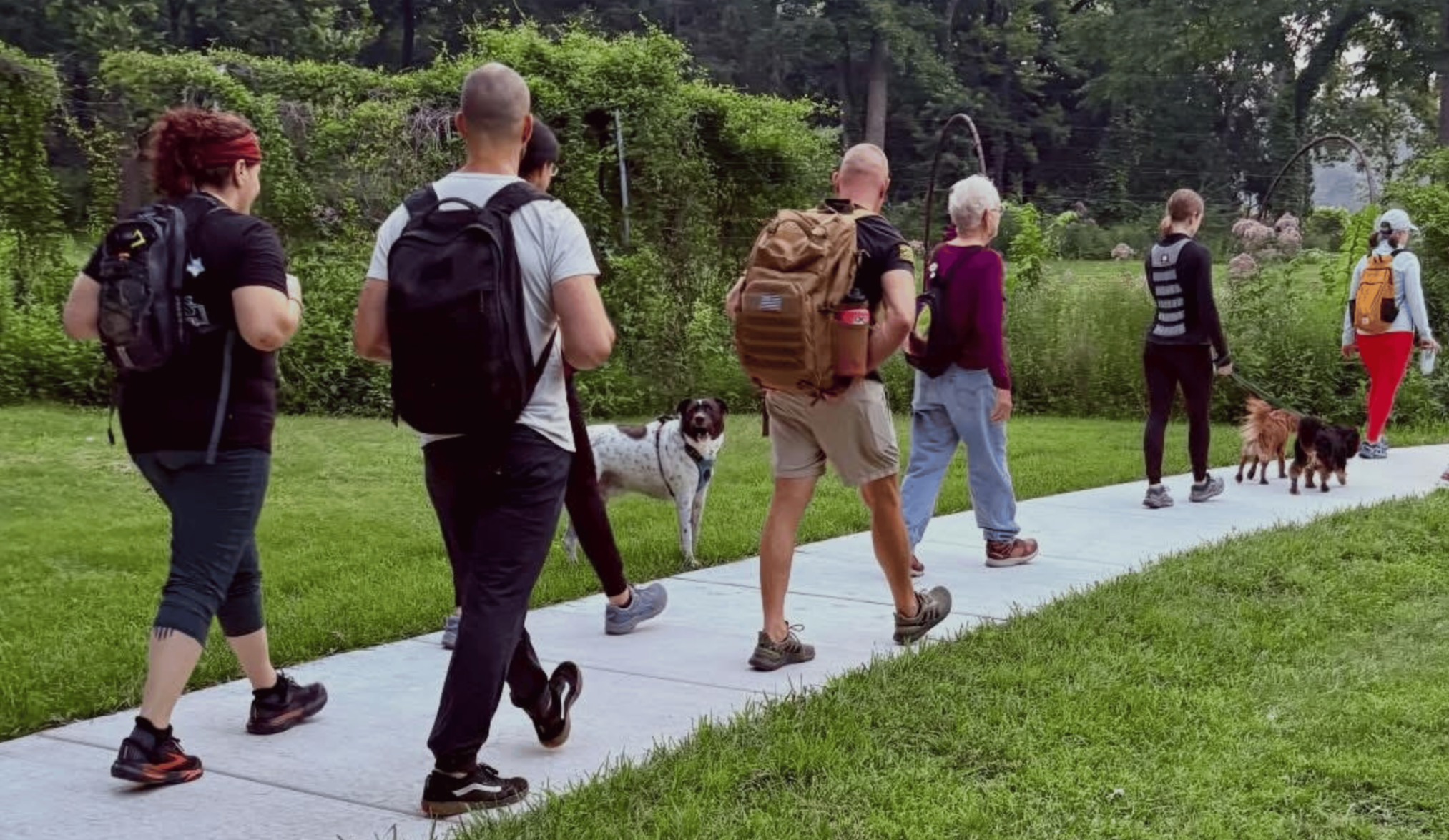 A group of six people walking along a sidewalk in a park with their dogs. They are wearing casual outdoor clothing and backpacks, surrounded by green trees and shrubs in the background.