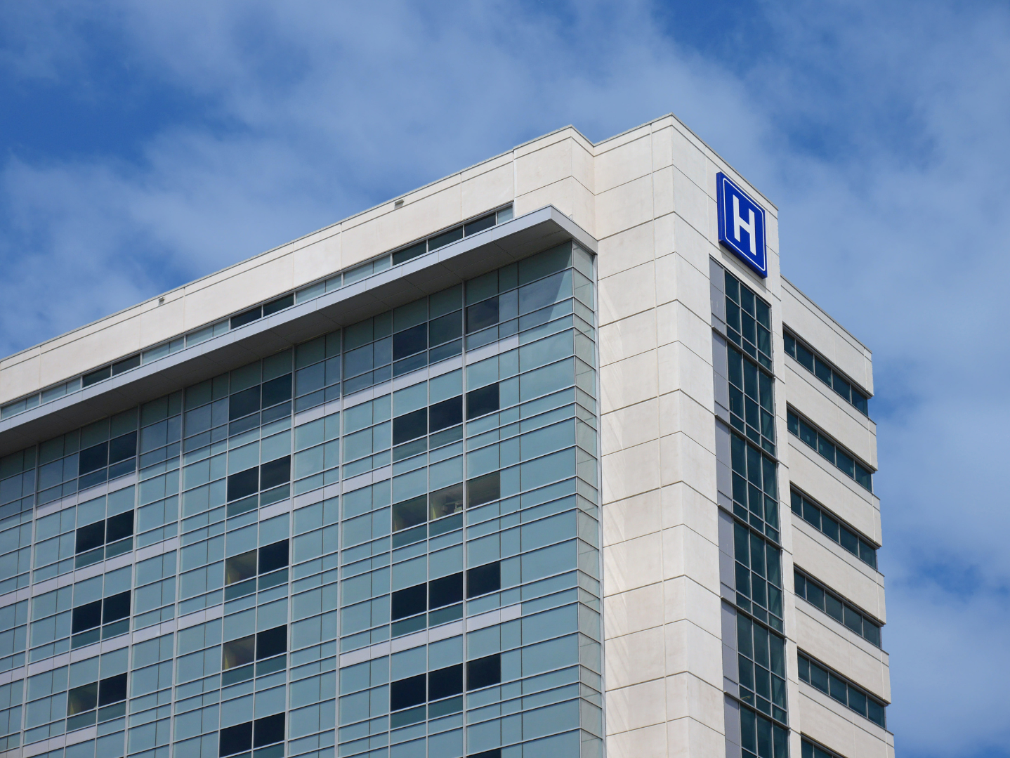 Modern hospital building with glass windows and a blue 'H' sign on the top corner, under a partly cloudy sky.