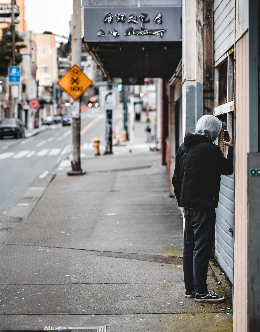 A person with a gray hoodie and black jacket standing on a city sidewalk, taking a photo through a window or door with a smartphone. The street has cars, traffic signs, and buildings in the background.