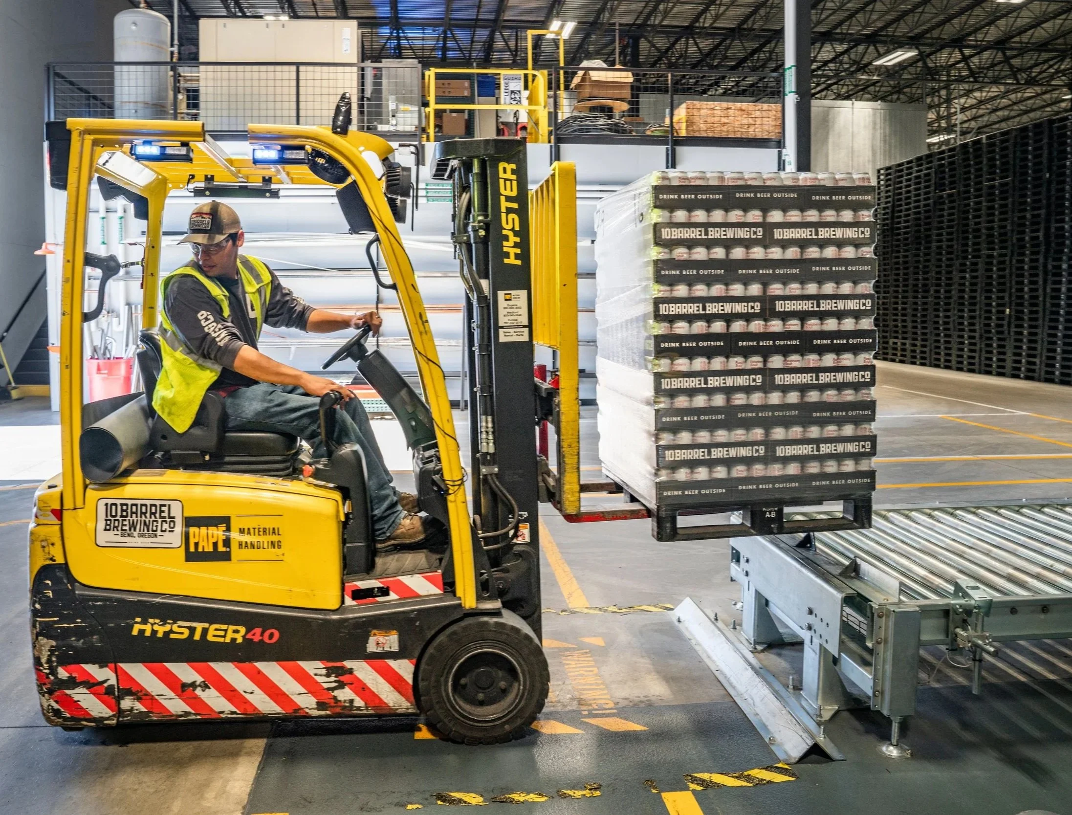 A worker operating a yellow forklift in a warehouse, moving a pallet stacked with cases of beer labeled "10 Barrel Brewing Co." and "Drink Beer Outside." The warehouse has visible metal shelves and industrial equipment in the background.