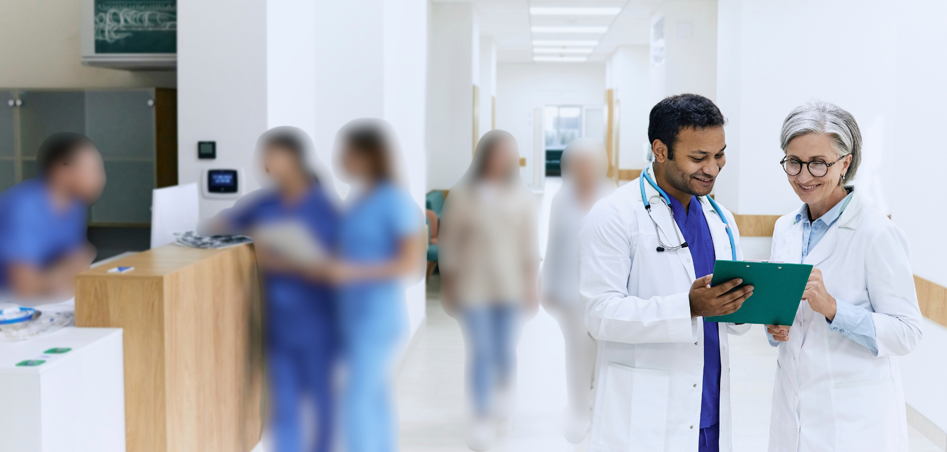 Two doctors, one male and one female, are standing together in a hospital corridor, looking at a clipboard and smiling. Several blurred hospital patients are in the background.
