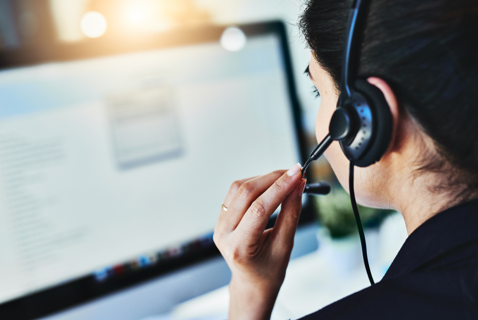A woman wearing a black headset with microphone, working on a computer, with a blurred screen in front of her.
