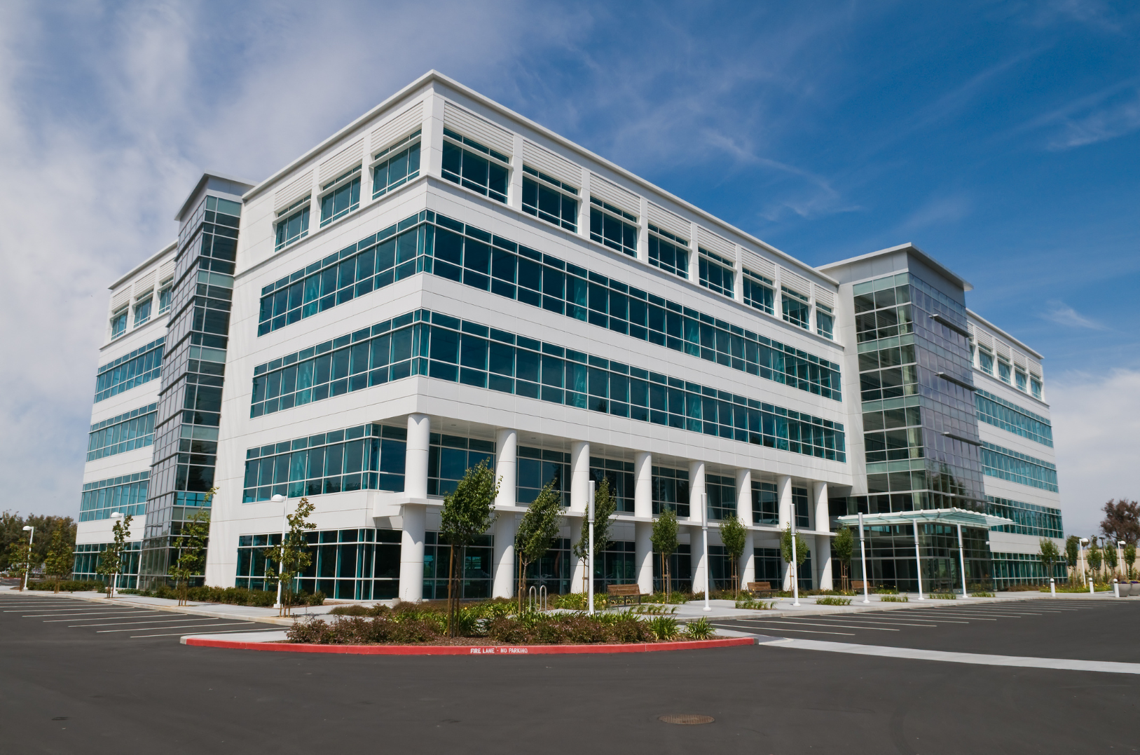 Modern multi-story office building with large glass windows, surrounded by a parking lot and landscaped trees, under a blue sky with some clouds.
