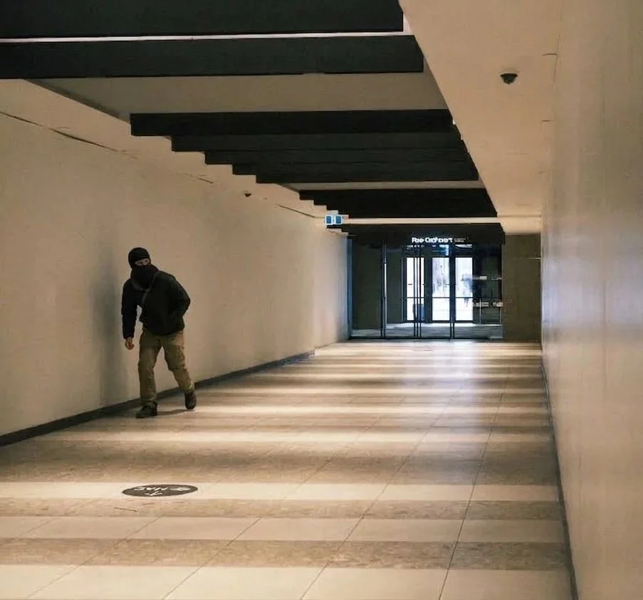 A man wearing a black hoodie, beige pants, and a ski mask walking in an empty, indoor corridor with beige walls and tiled floor, near glass doors at the end.