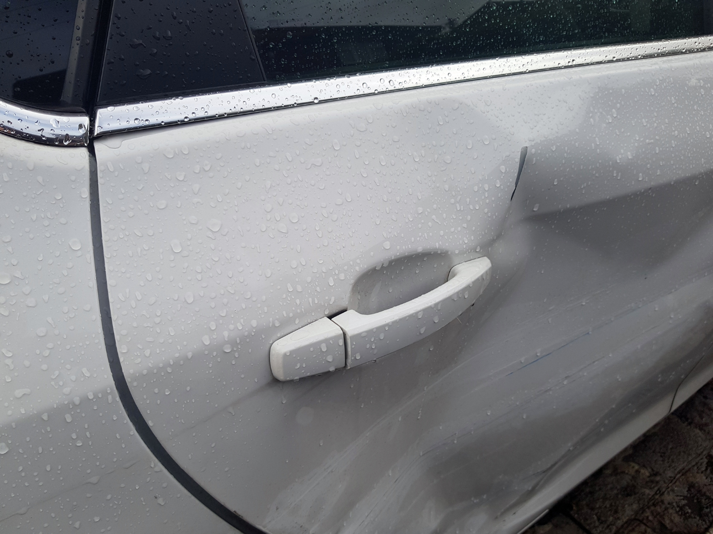 Close-up of a silver car door with a large dent and scratches, with raindrops on the surface and a white door handle.
