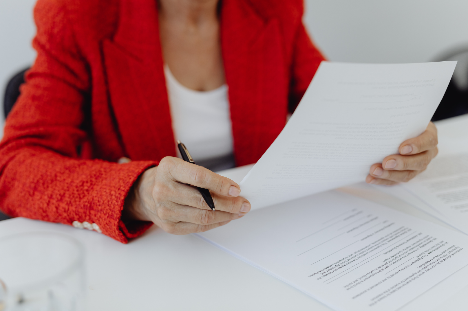 A woman in a red blazer sitting at a desk, holding a pen and reviewing a document.