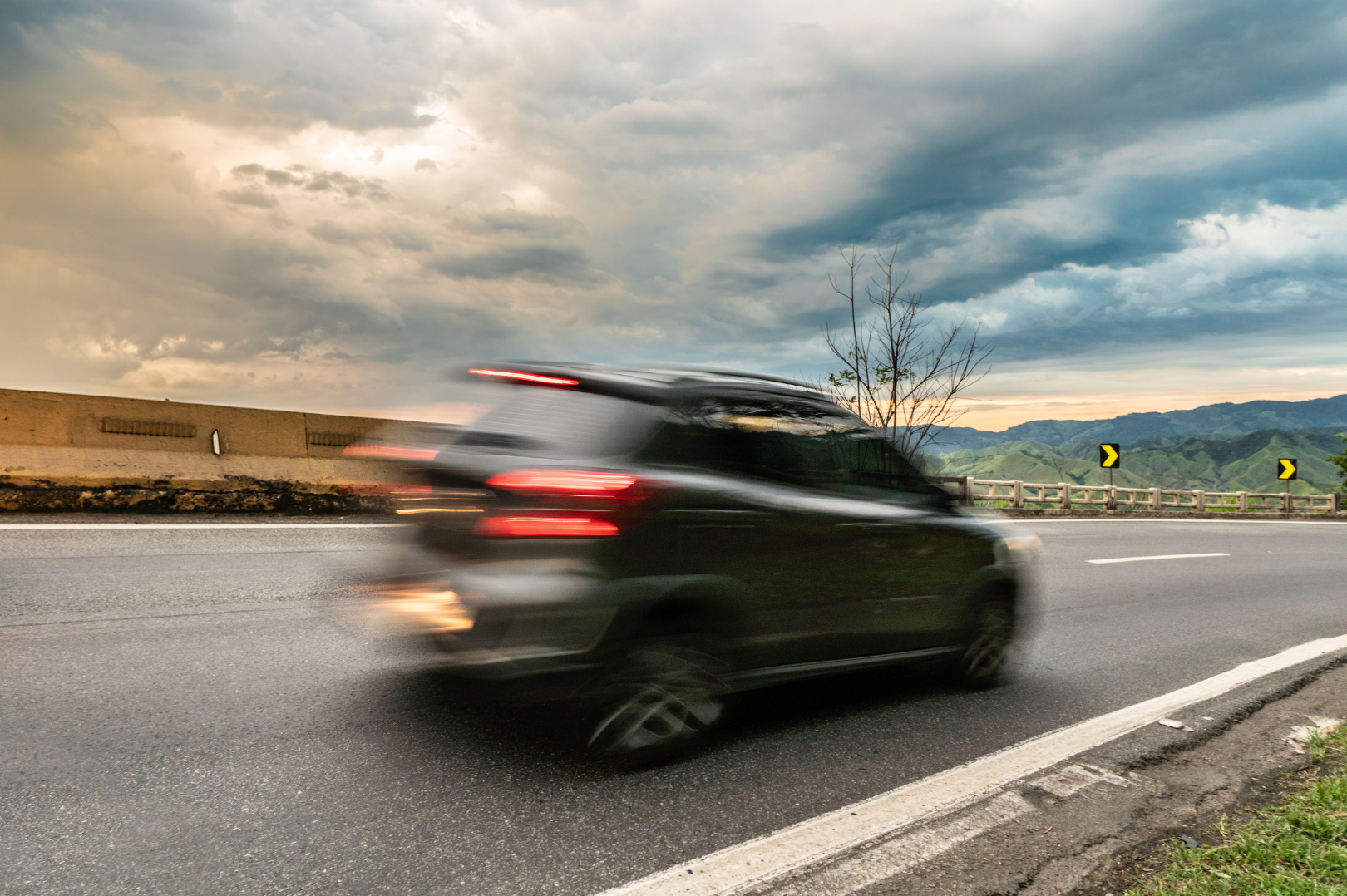 A black car moving at high speed on a highway with a blurred motion effect, set against a backdrop of a cloudy sky, mountains, and a roadside guardrail.