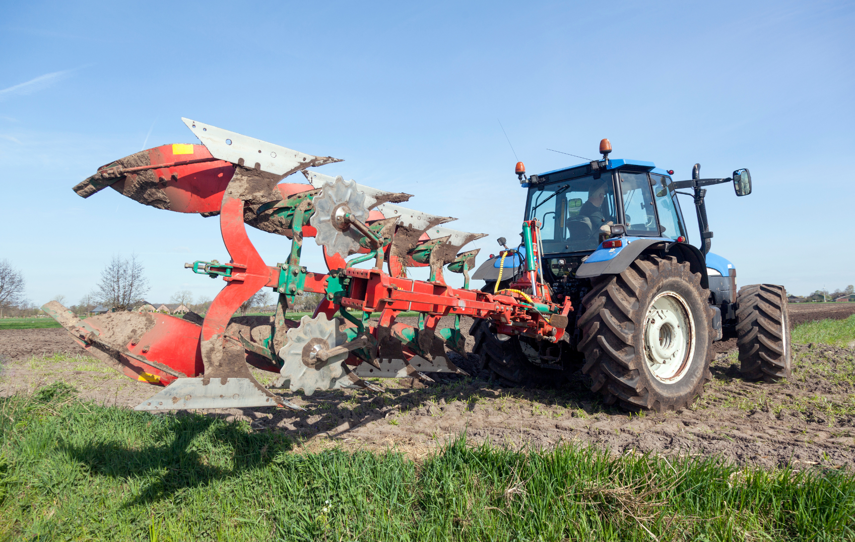 Farm tractor pulling a red plow in a field under a clear blue sky.