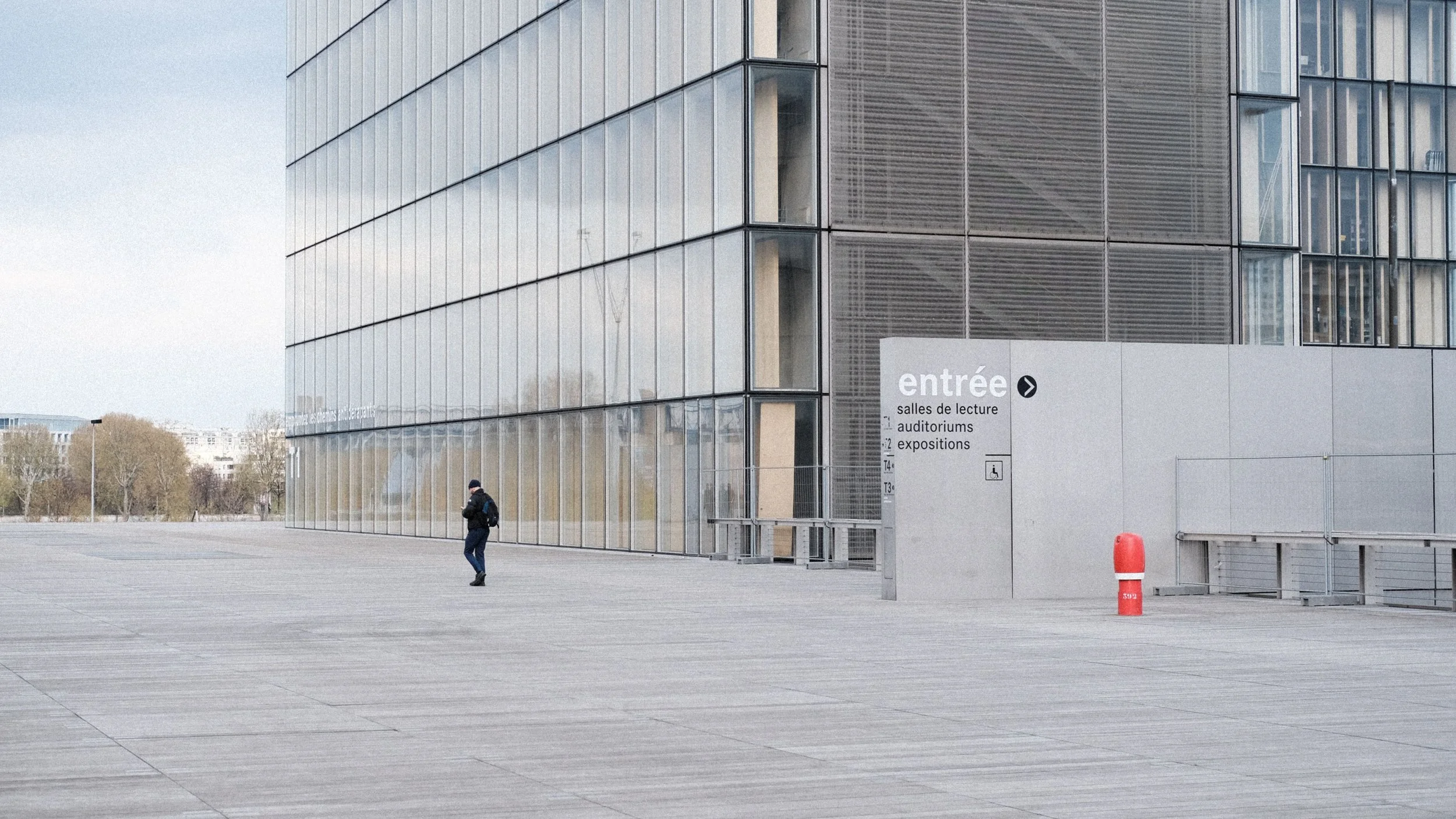 Modern glass building with a sign indicating entrance. A person in dark clothing and backpack is walking outside on a large, empty paved area.