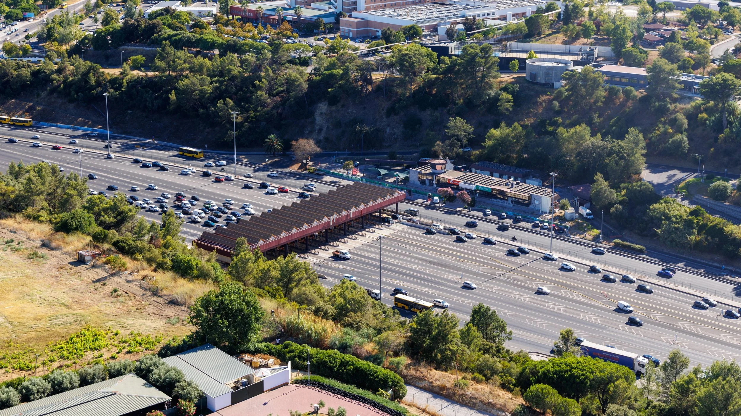 Aerial view of a toll plaza on a highway, with many cars waiting in line to get through, and many leaving the plaza. There are green trees and residential houses on both sides of the highway.