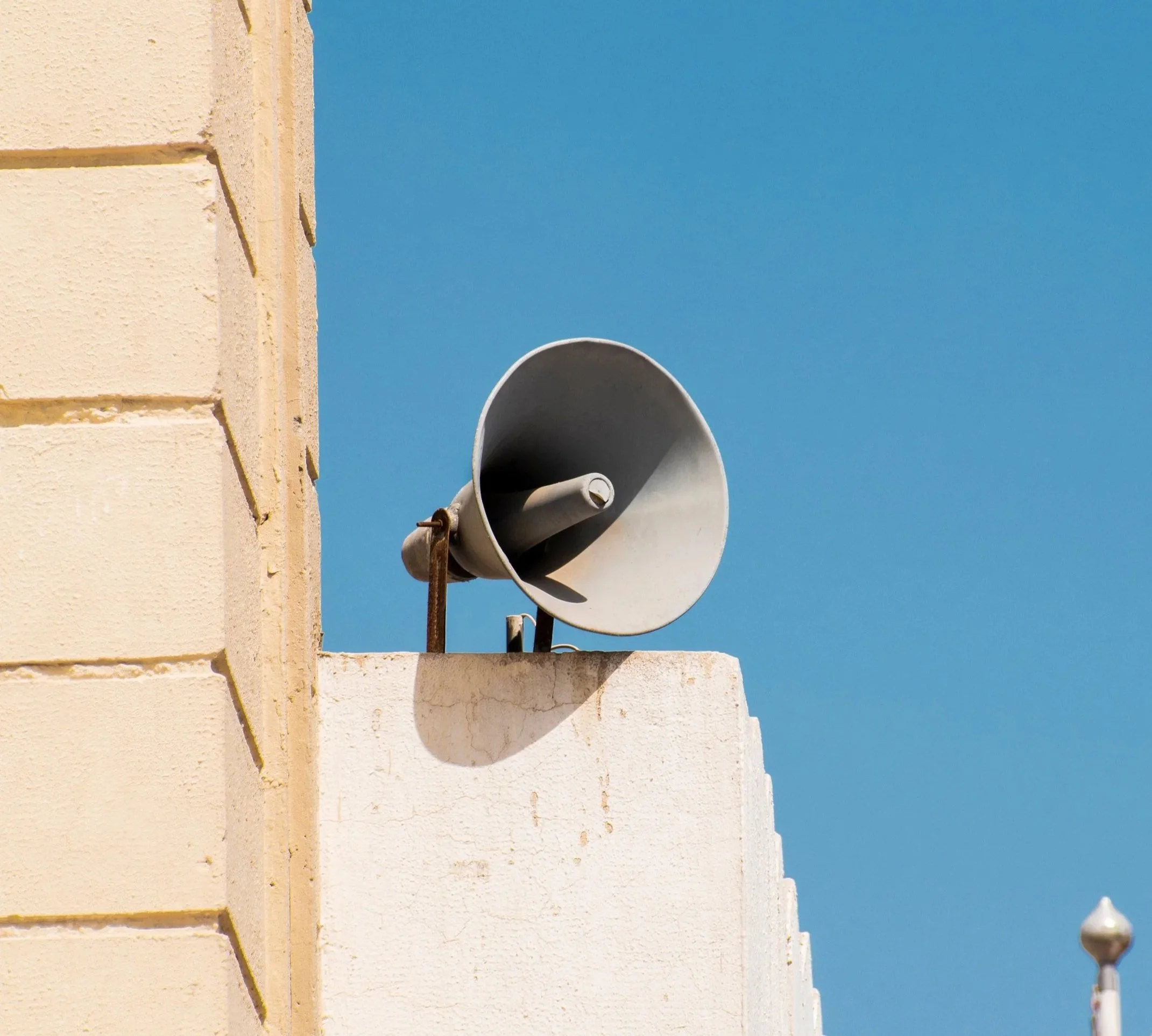 A white building corner with a large white megaphone mounted on the wall, against a clear blue sky.