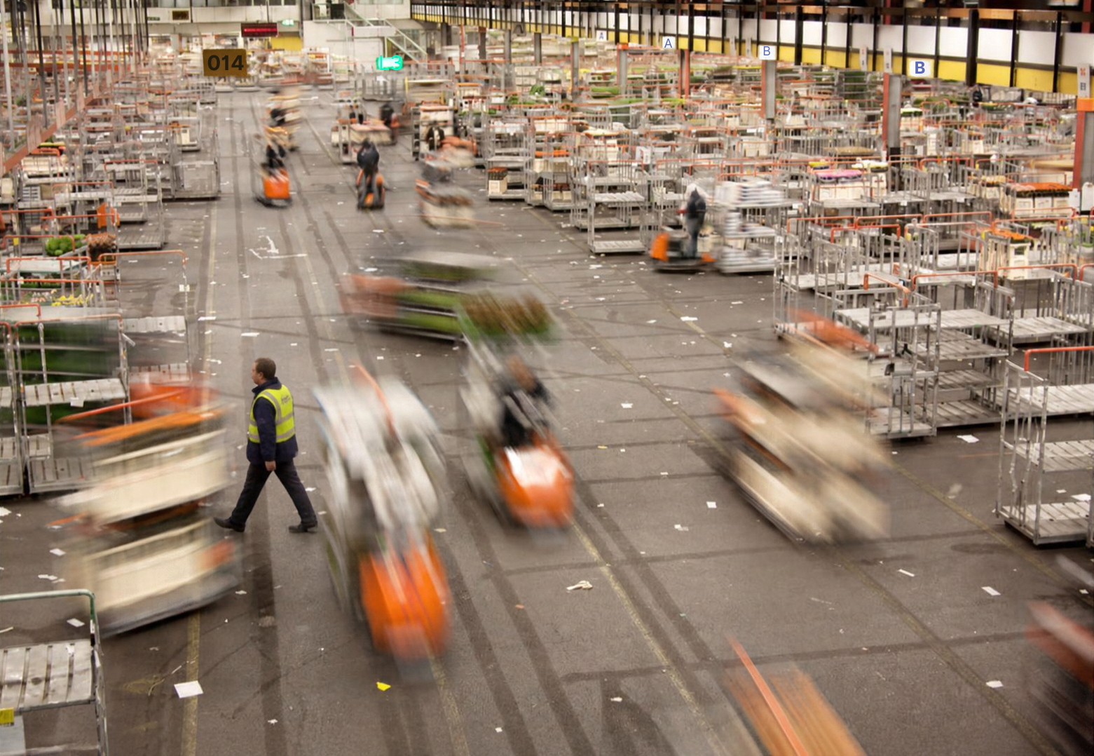 Inside a warehouse, multiple workers operate forklifts and carts, with some in motion creating a blurred effect, while a person in a yellow safety vest walks across the floor amid empty shelving units.