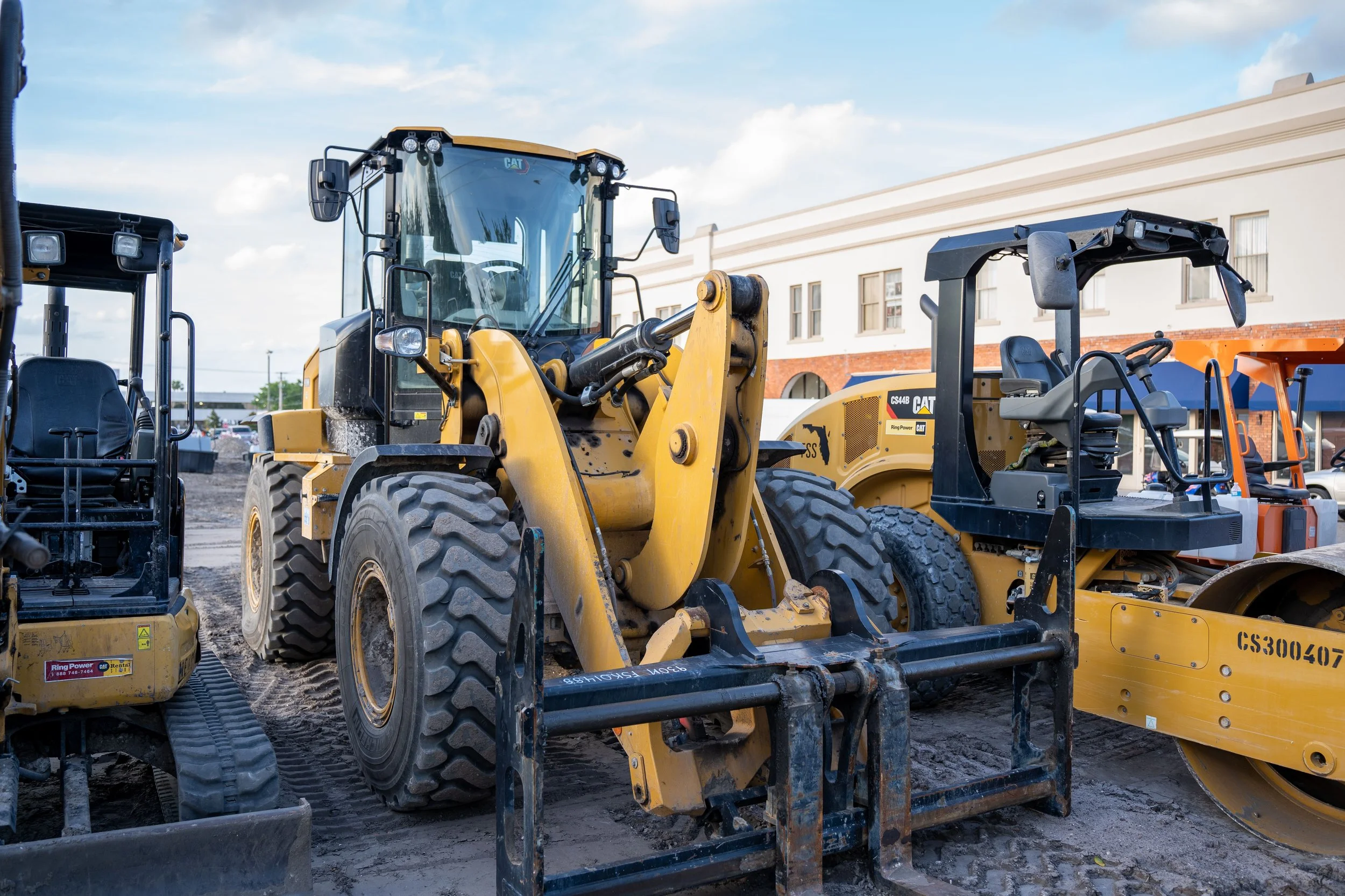 Multiple construction vehicles, including yellow excavators and a loader, are parked on a construction site with a building in the background under a partly cloudy sky.