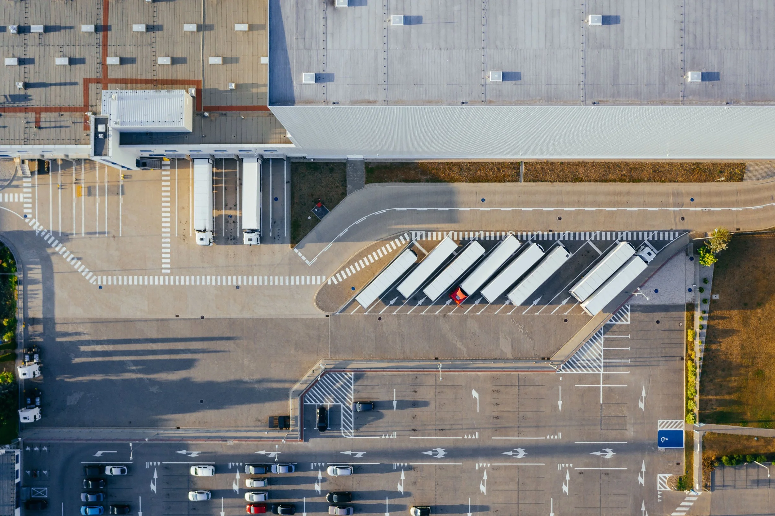 An aerial view of a transportation & logistics lot with semi trucks parked alongside the building, and 2 trailers parked in loading docks.