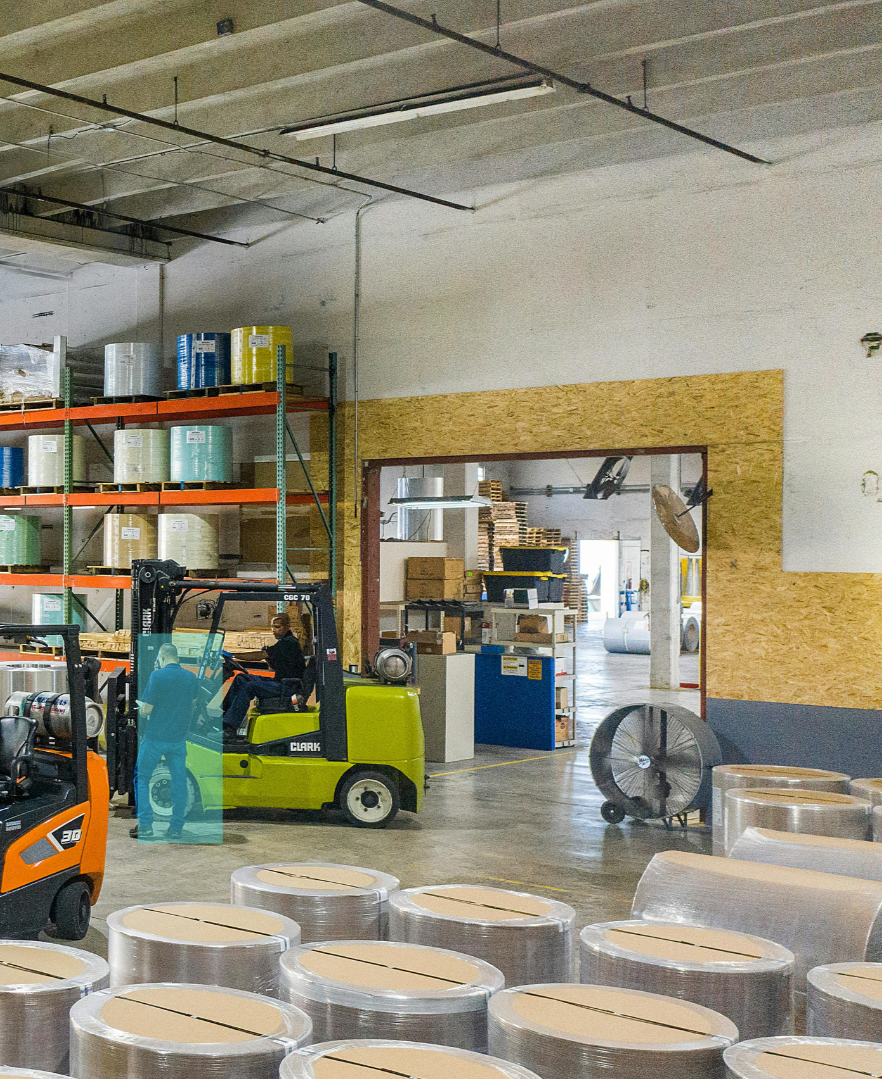 Warehouse interior with forklifts, large rolls of plastic or paper, and shelves with various materials.
