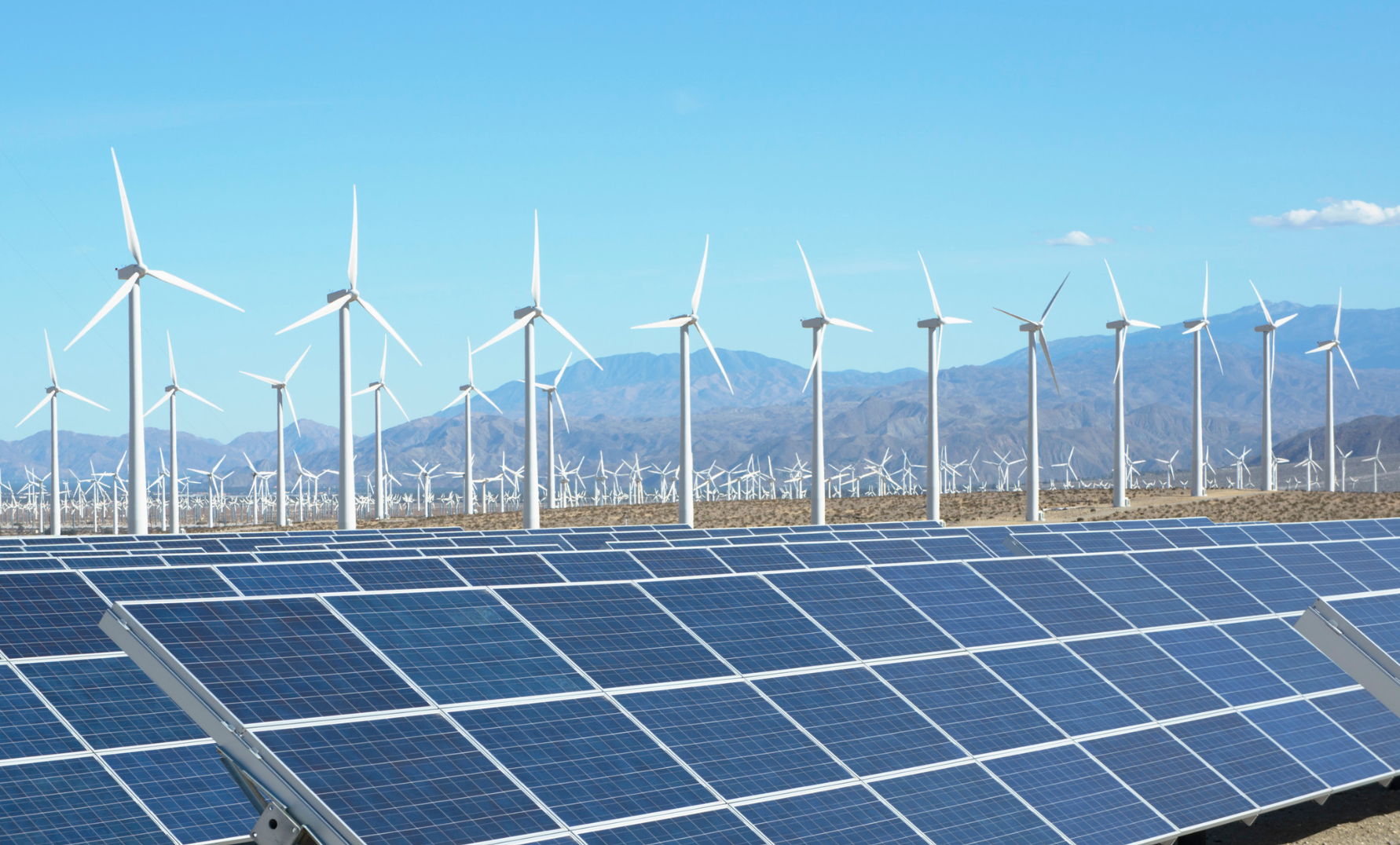 A large solar farm with solar panels in the foreground and numerous wind turbines in the background, set against a mountainous landscape under a clear blue sky.