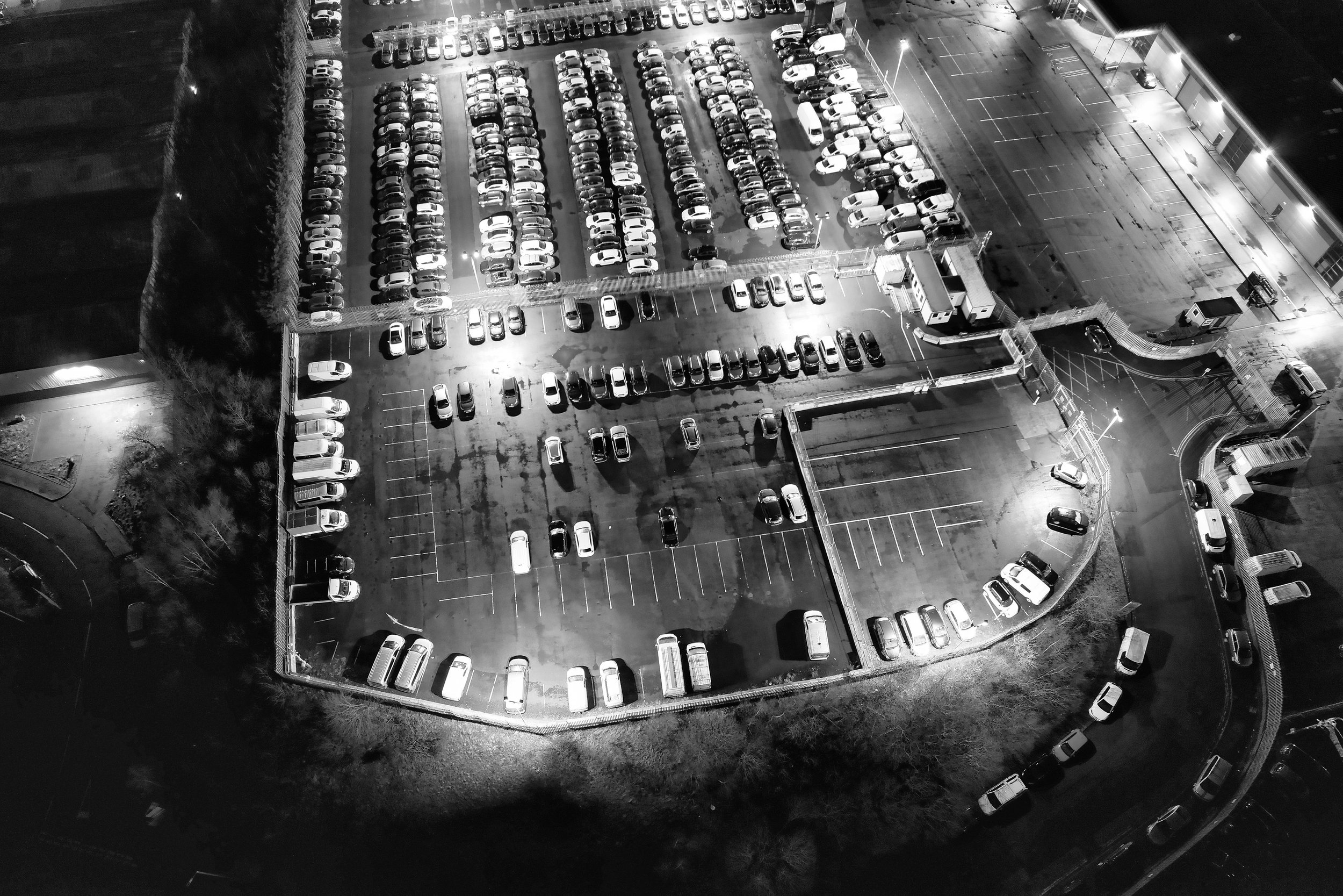 A nighttime aerial view of a parking lot filled with numerous parked cars, surrounded by fencing and illuminated by streetlights, with some empty parking spaces visible.