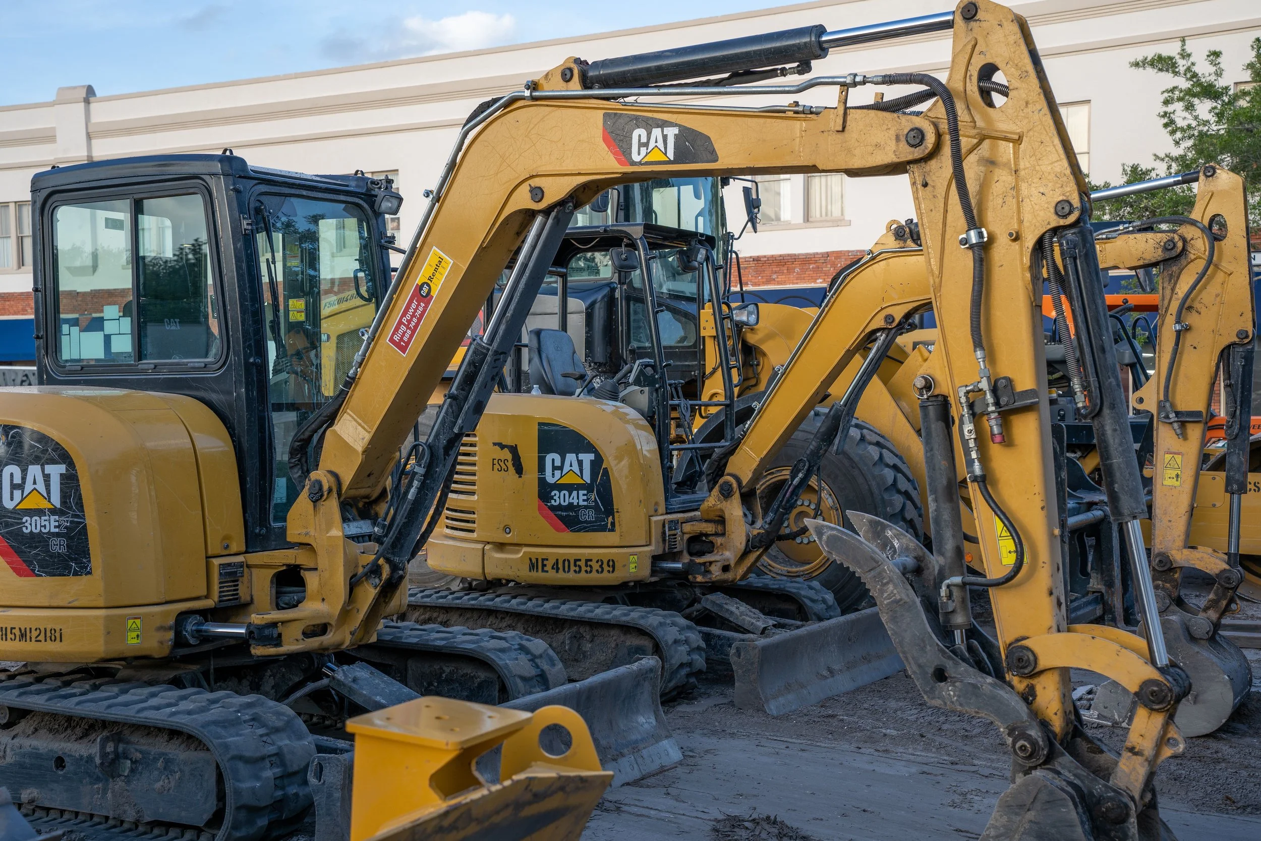 A large Caterpillar construction excavator parked outdoors near a building, with a backhoe attachment, mirrors, and hydraulic hoses visible.