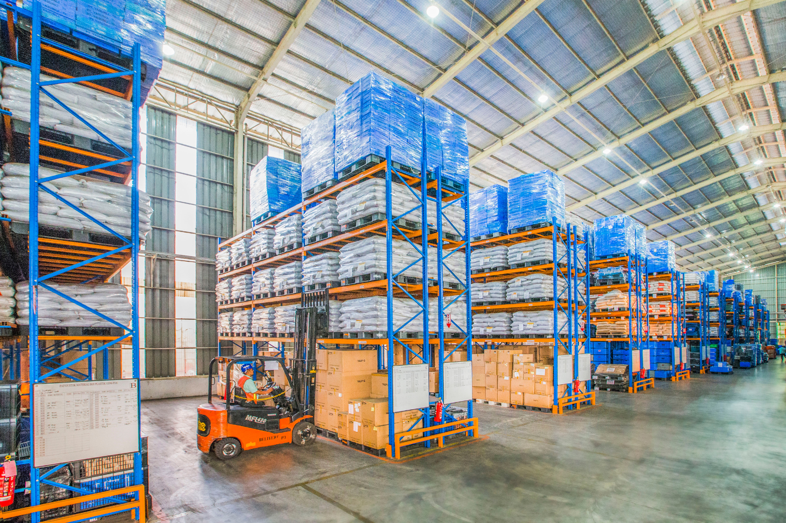 Inside a large warehouse with high ceilings, blue and orange storage racks filled with bags and boxes. A worker operating a forklift is moving boxes among the shelves.