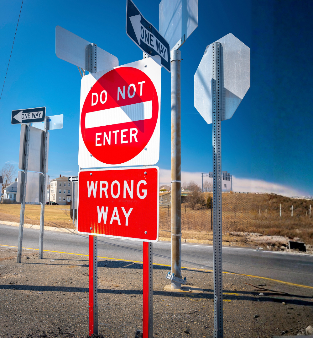Collection of traffic signs including a red and white 'Do Not Enter' sign with a smaller red sign below labeled 'Wrong Way' and one-way signs in the background on a street with houses and open land.