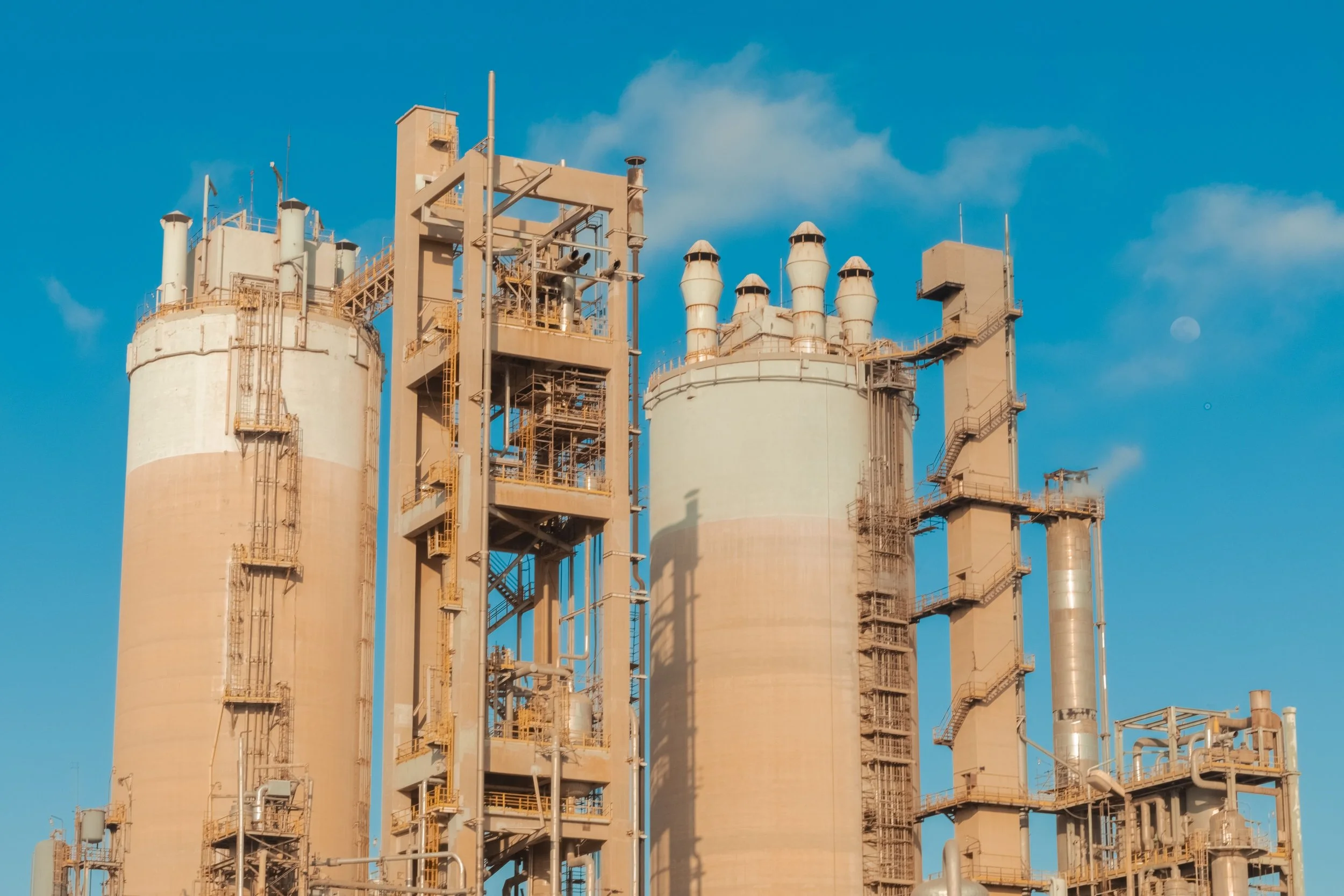 Industrial factory with large beige storage tanks, pipes, stairs, and vents against a bright blue sky.