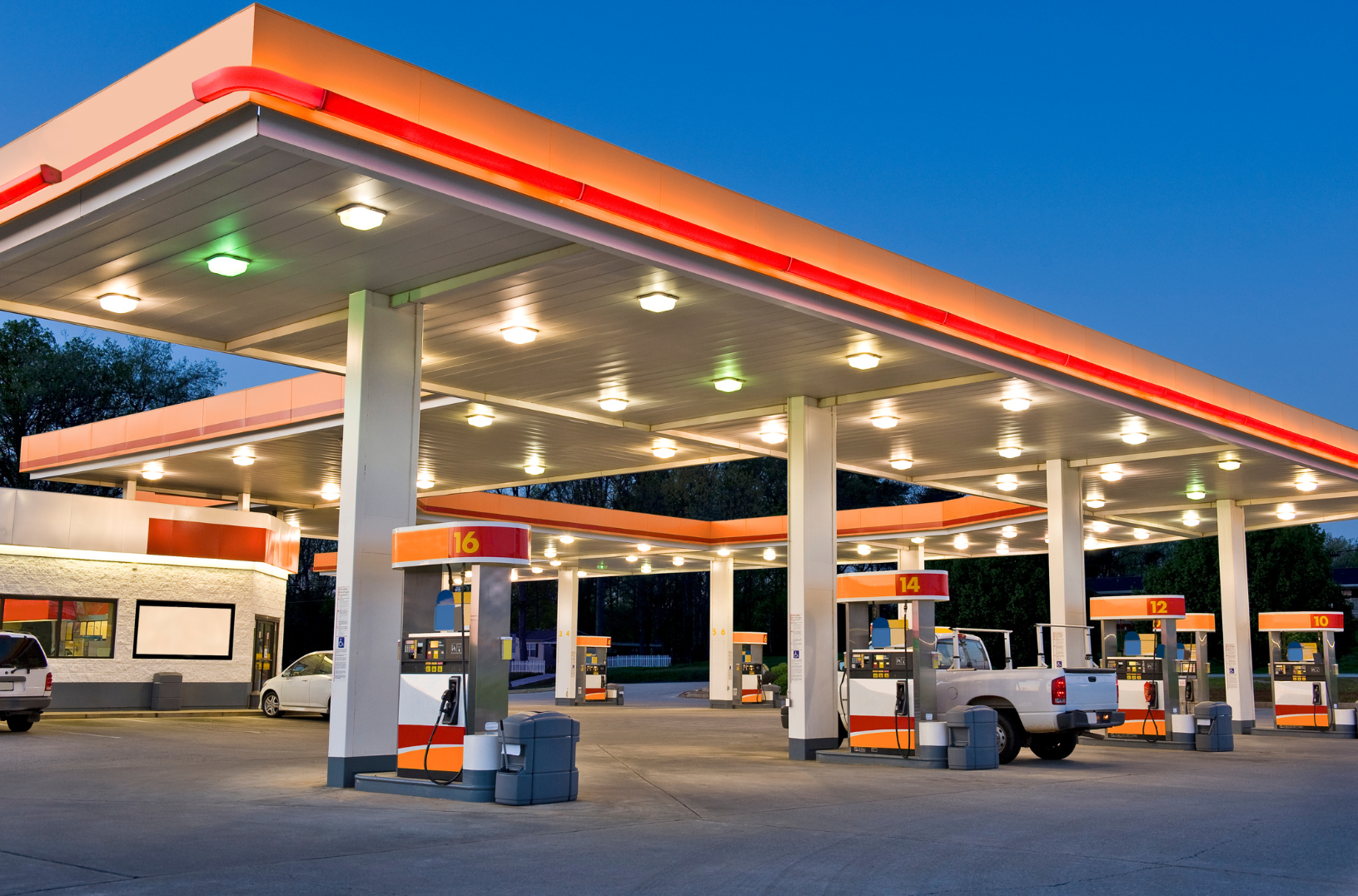 Gas station with multiple fuel pumps and cars parked, illuminated by bright lights at dusk or early evening.