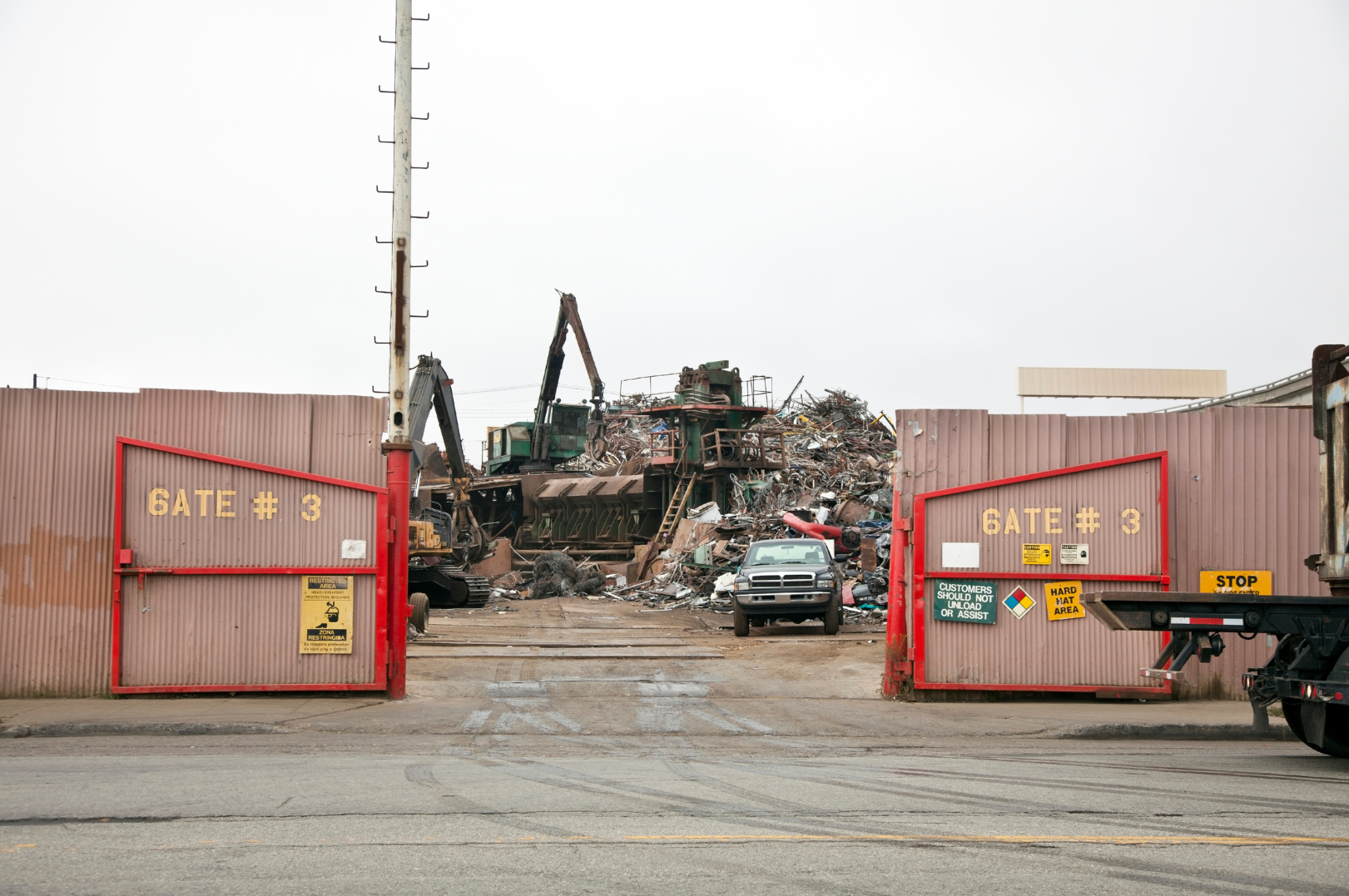 View of an industrial scrapyard behind a red gate with the sign 'Gate # 3' and caution notices, with heavy machinery and a large pile of scrap metal and vehicles in the background.