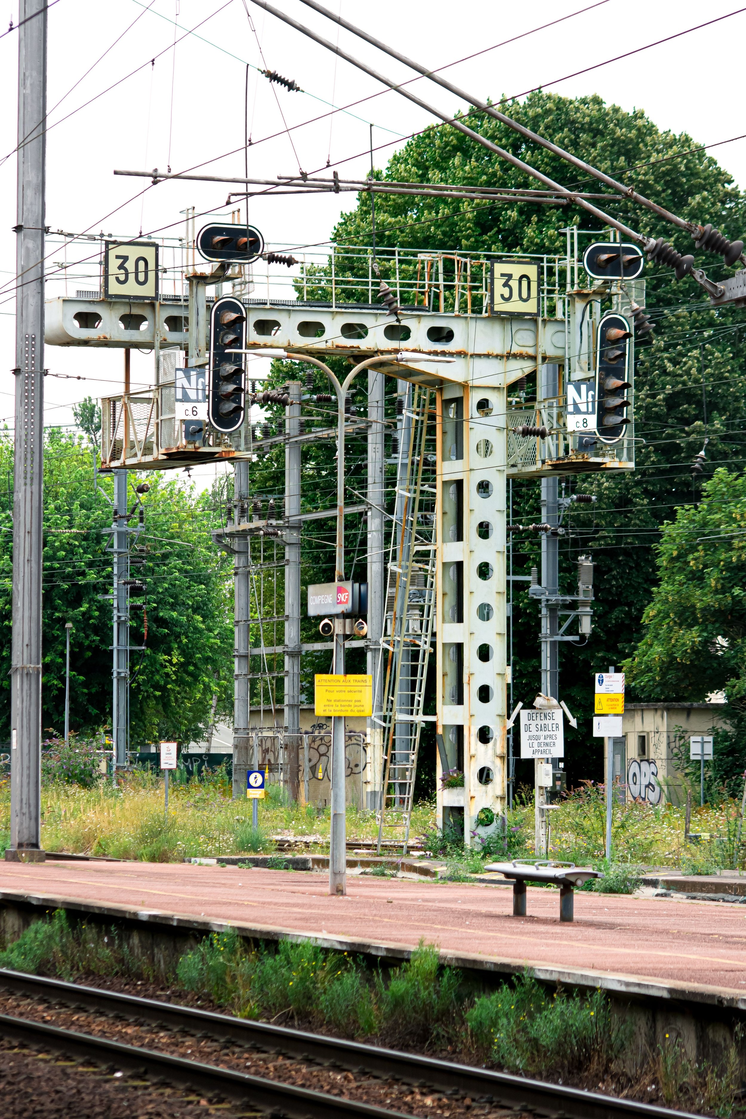 A train station platform with overgrown grass and graffiti, surrounded by trees, with railway tracks in the foreground, and various electrical infrastructure and signs.