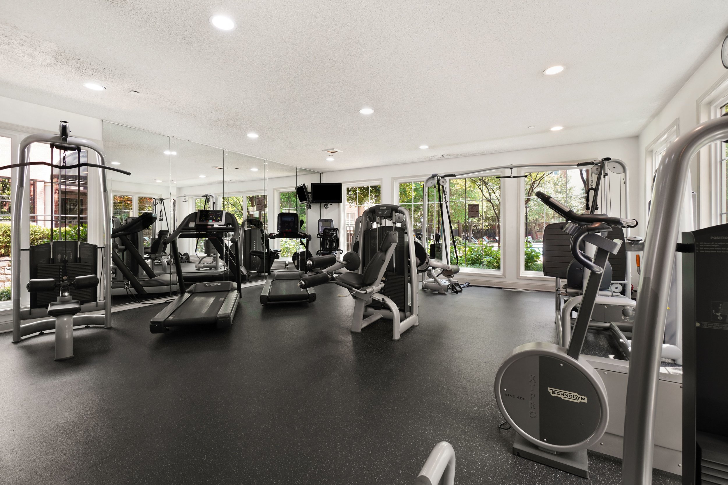 Interior view of a modern gym with exercise equipment including treadmills, ellipticals, and weight machines, surrounded by large windows and a mirrored wall.