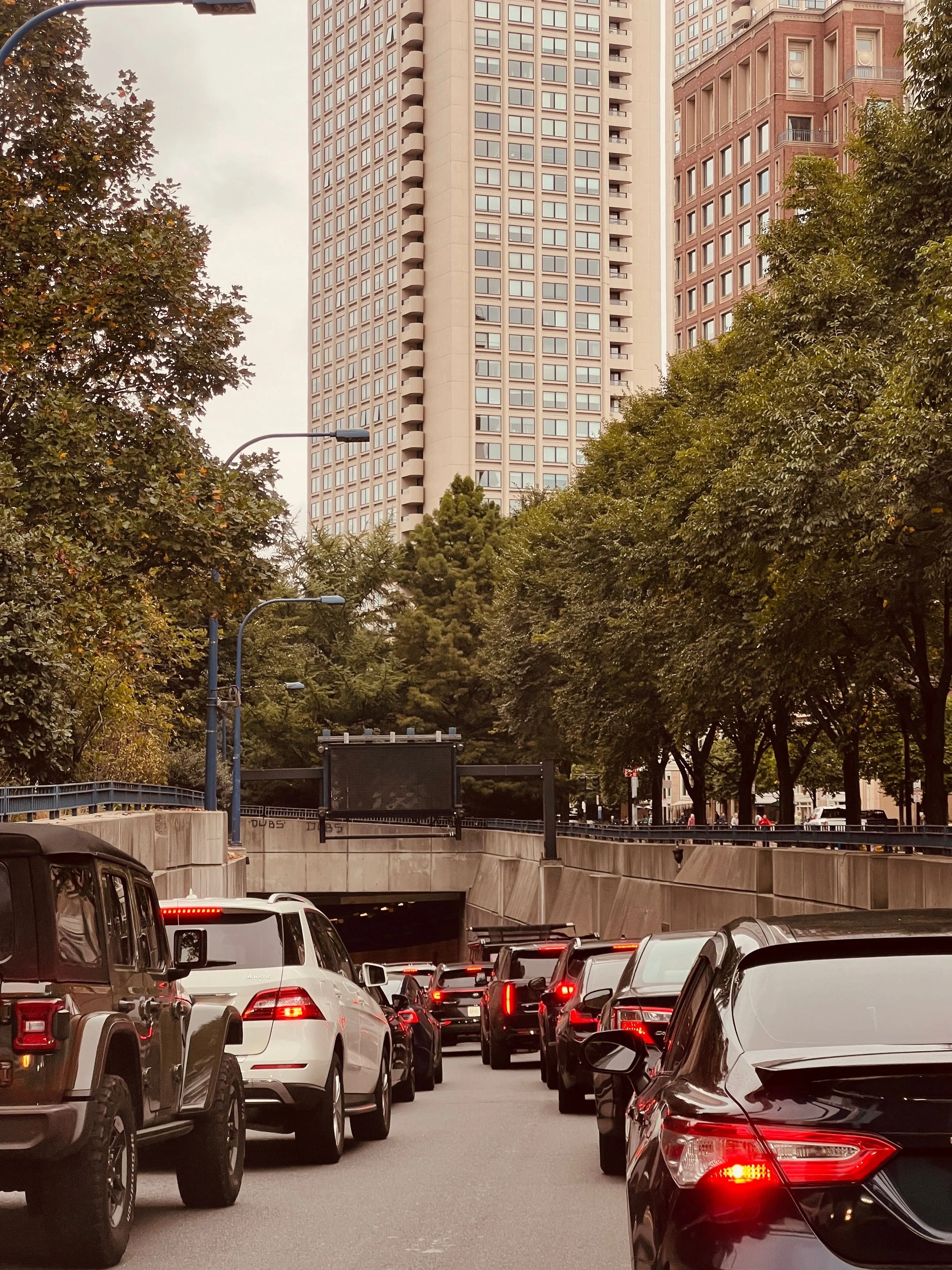 A street scene with a traffic jam of cars, some with brake lights on, in front of tall buildings and trees.