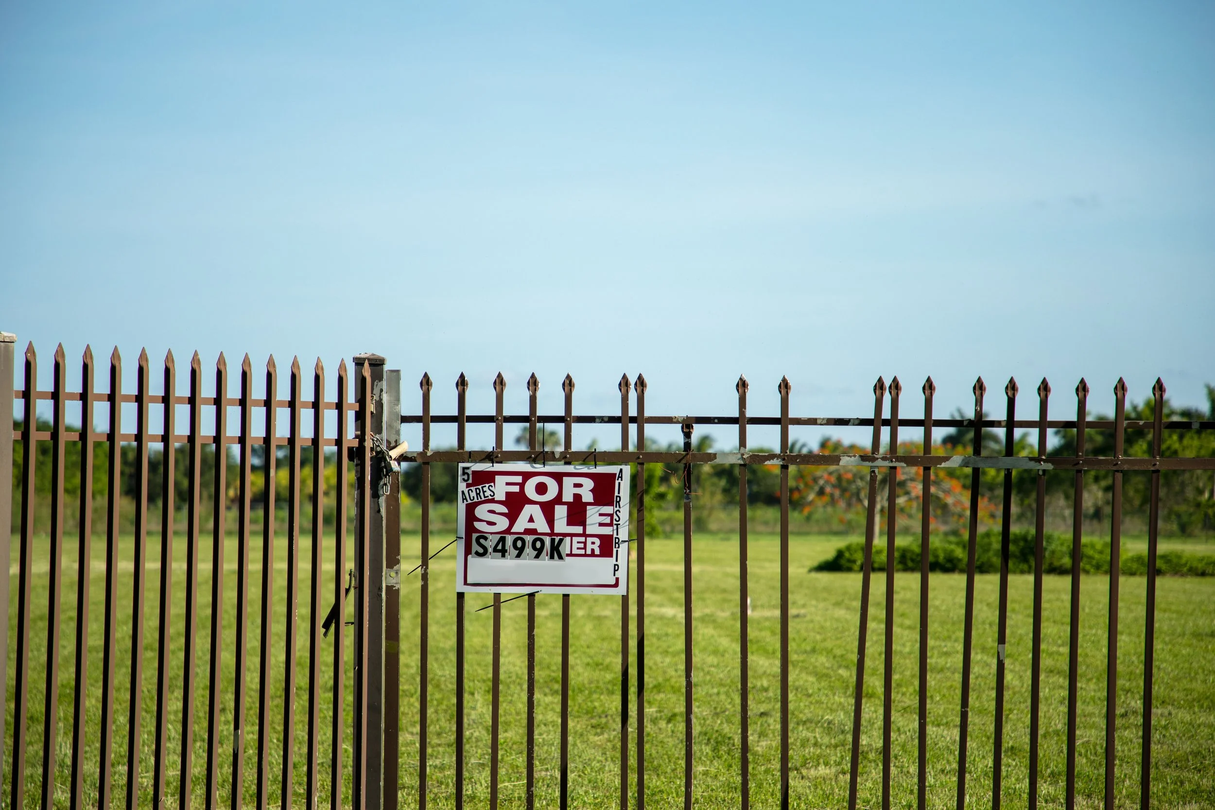 A metal fence with a 'For Sale' sign attached, advertising 5 acres of land for $499,000, with a clear sky and greenery in the background.