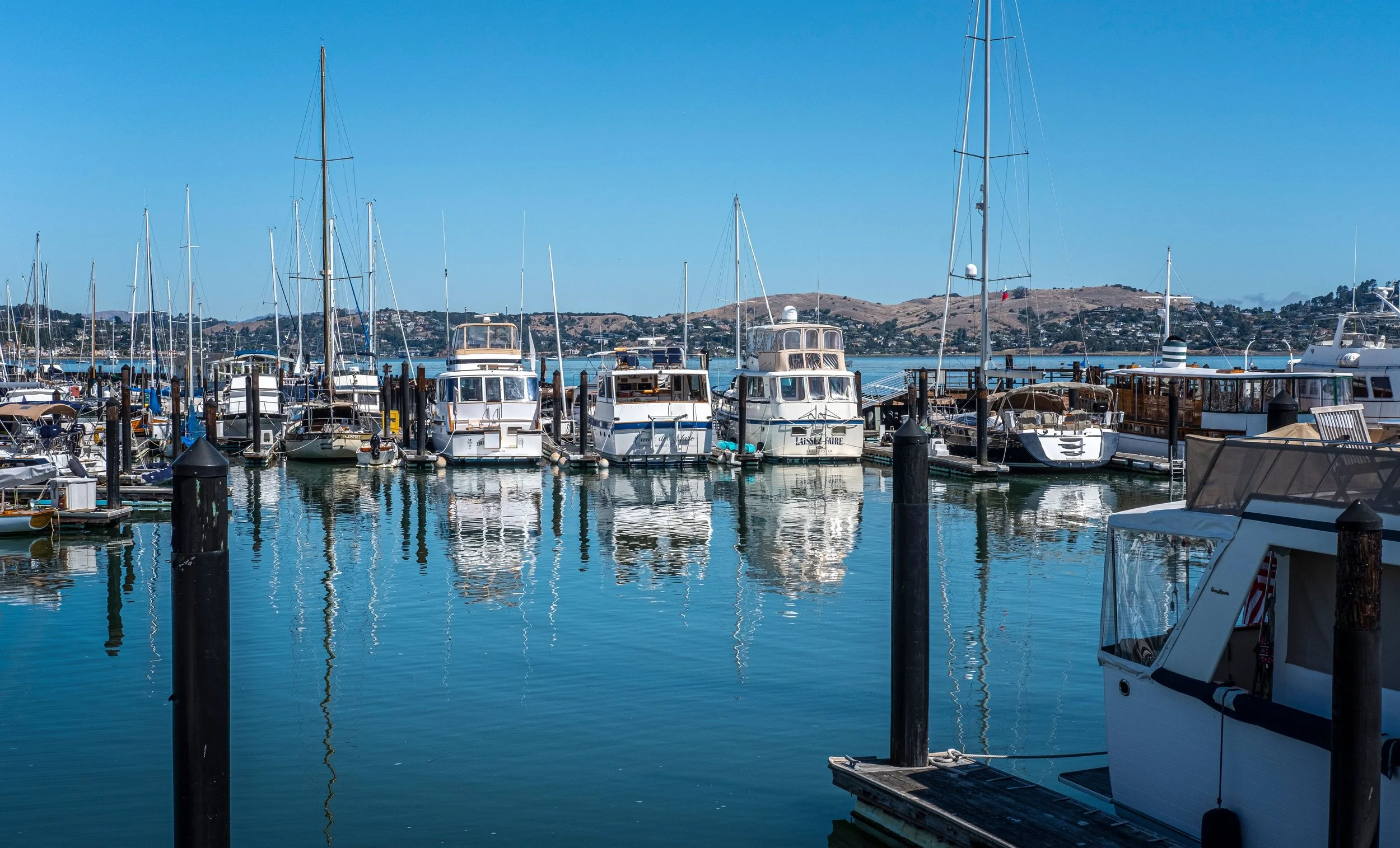 A marina with numerous sailboats and yachts docked, with hills and houses in the background under a clear blue sky.