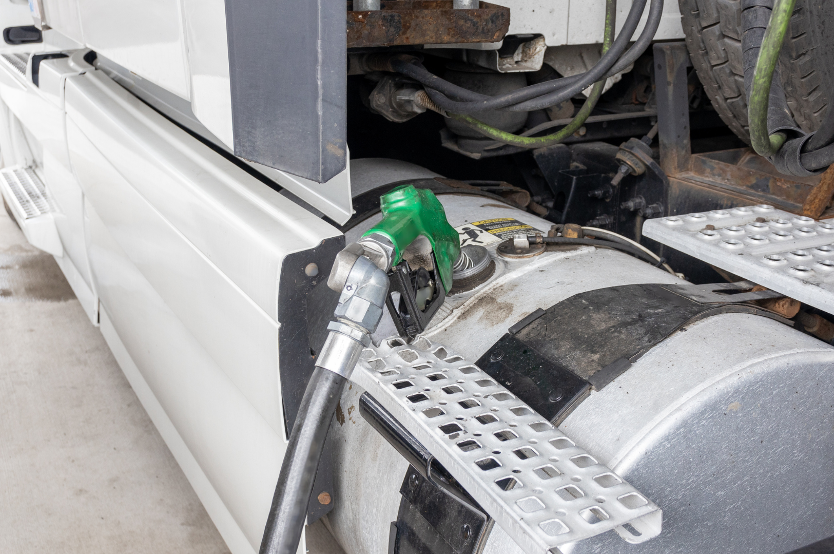 Close-up of a fuel nozzle filling a semi-truck fuel tank with fuel. The fuel nozzle has a green handle, and the truck has a metallic fuel tank with black patches and a step ladder attached.