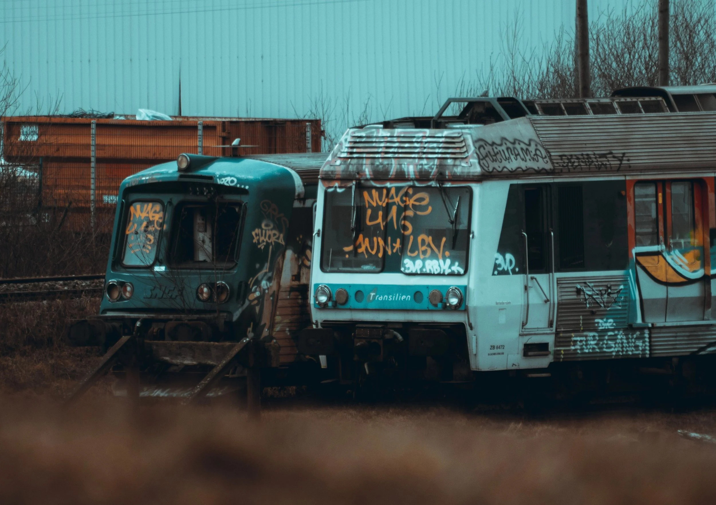 An abandoned, graffiti-covered train car on train tracks with a greenish building and fence in the background.