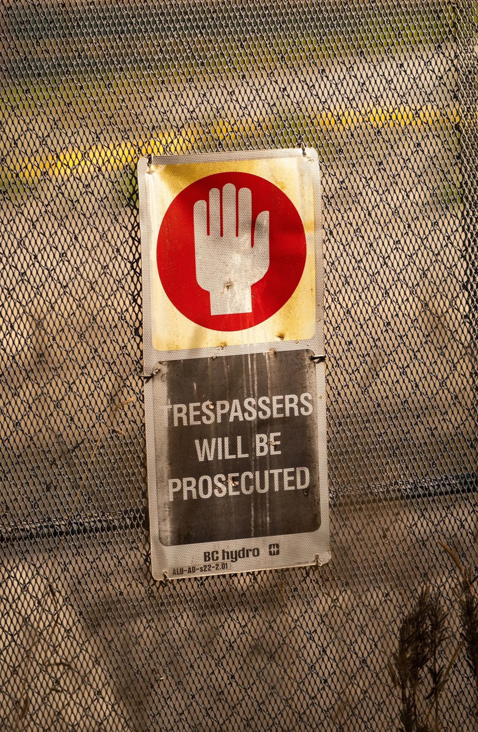 A security fence with a warning sign featuring a red circle with a white hand and text that states trespassers will be prosecuted.