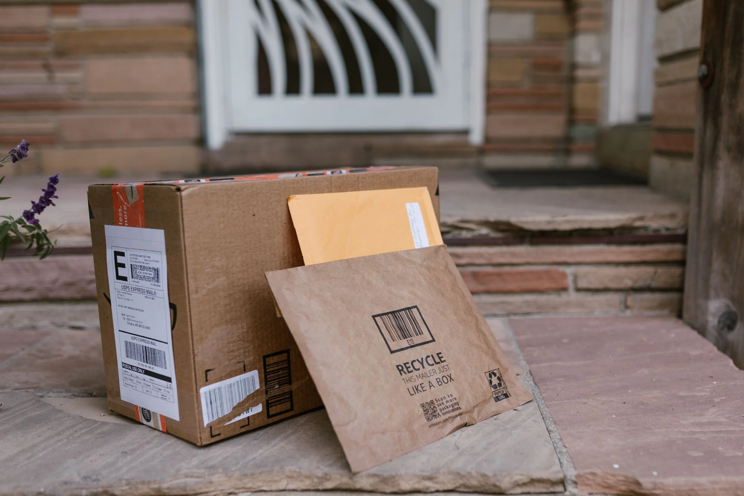 A cardboard USPS shipping box and a paper recycling bag resting on a front porch made of brick. The box has shipping labels and barcodes, while the paper bag has recycling instructions printed on it.