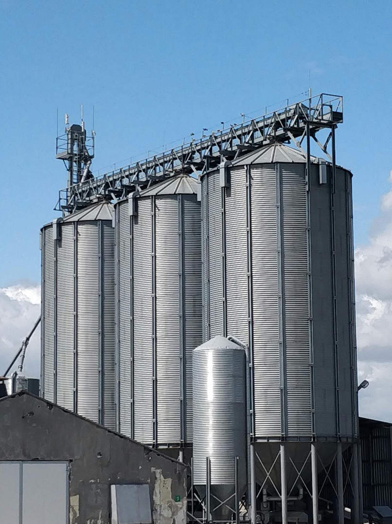 Multiple silver grain silos on a farm, with a metal skywalk at the top connecting them and a small building in front with a cloud-filled sky in the background.